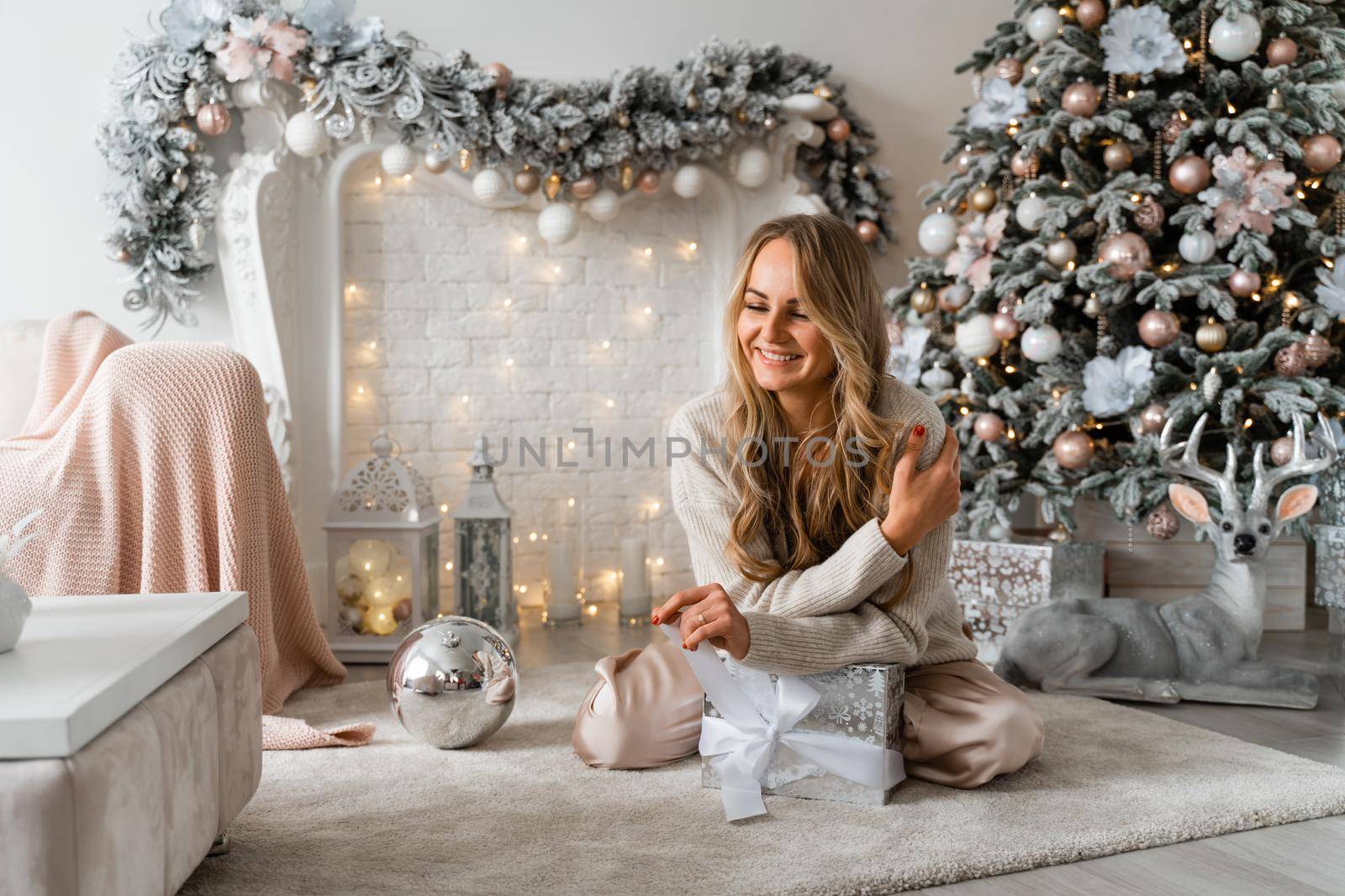 Happy blonde beauty enjoys Christmas Under the Christmas tree in a cozy home, holding a box with a gift