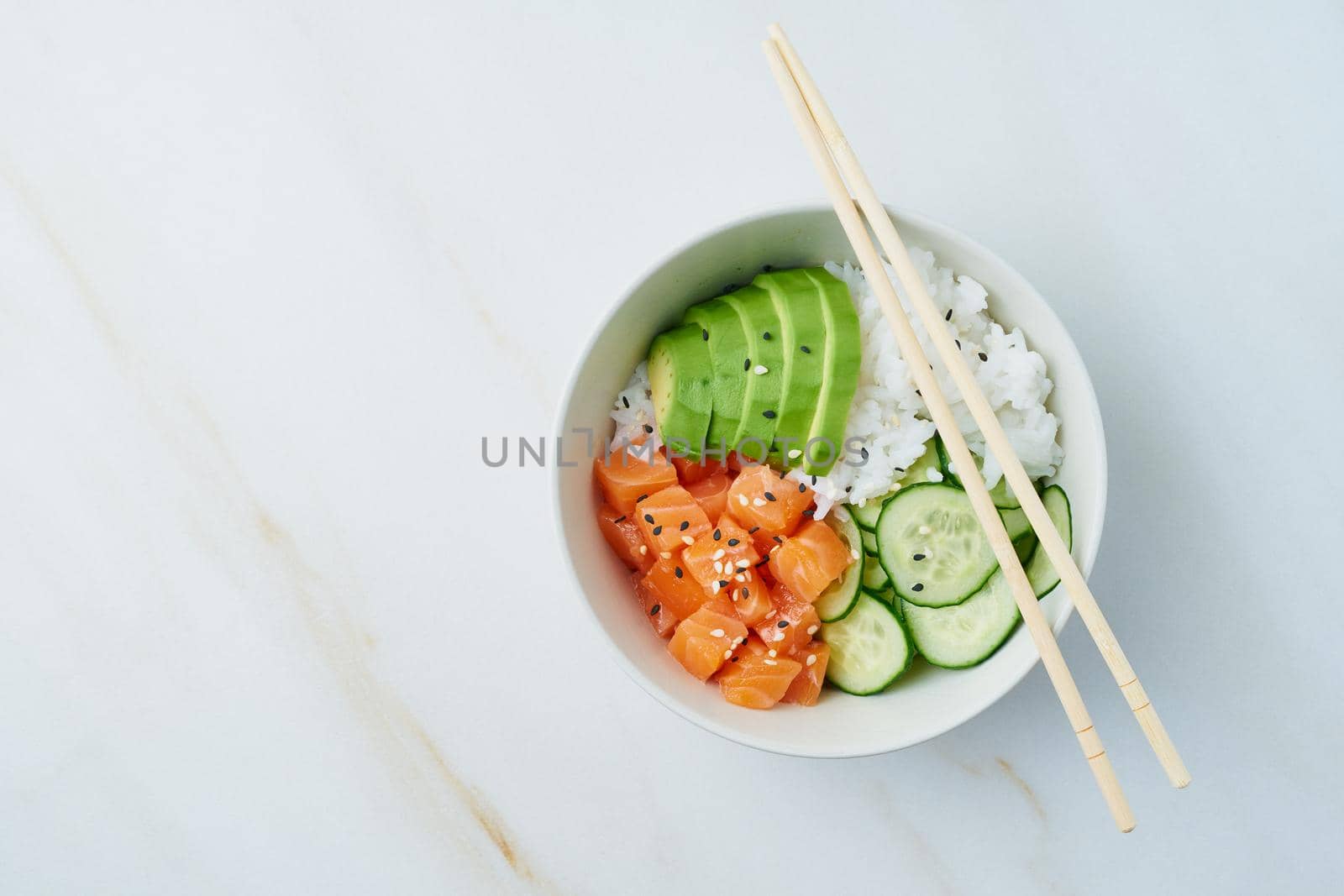 Salmon poke bowl with fresh fish, rice, cucumber, avocado with black and white sesame. White marble table. Food concept.