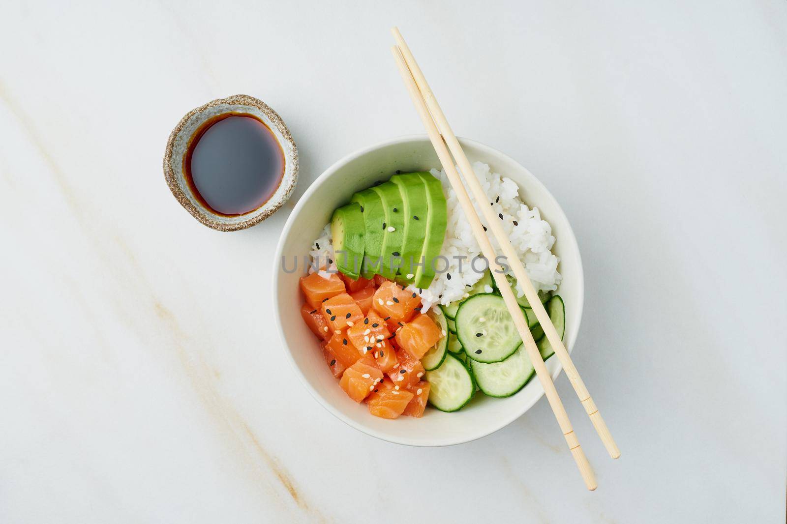 Salmon poke bowl with fresh fish, rice, cucumber, avocado with black and white sesame. White marble table. Food concept.