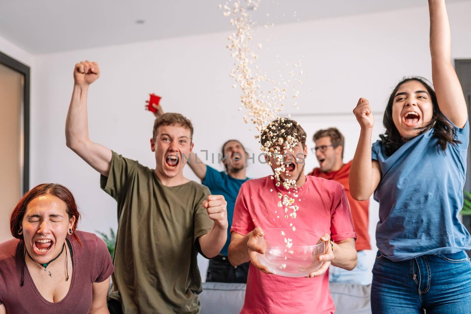 friends jumping for joy, throwing popcorn in the air after their team's victory. group of young people watching football at home. leisure concept. happy and cheerful. natural light in the living room at home. trumpet