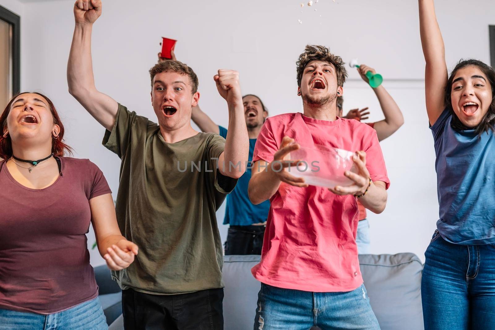 friends jumping for joy, throwing popcorn in the air after their team's victory. group of young people watching football at home. leisure concept. happy and cheerful. natural light in the living room at home. trumpet