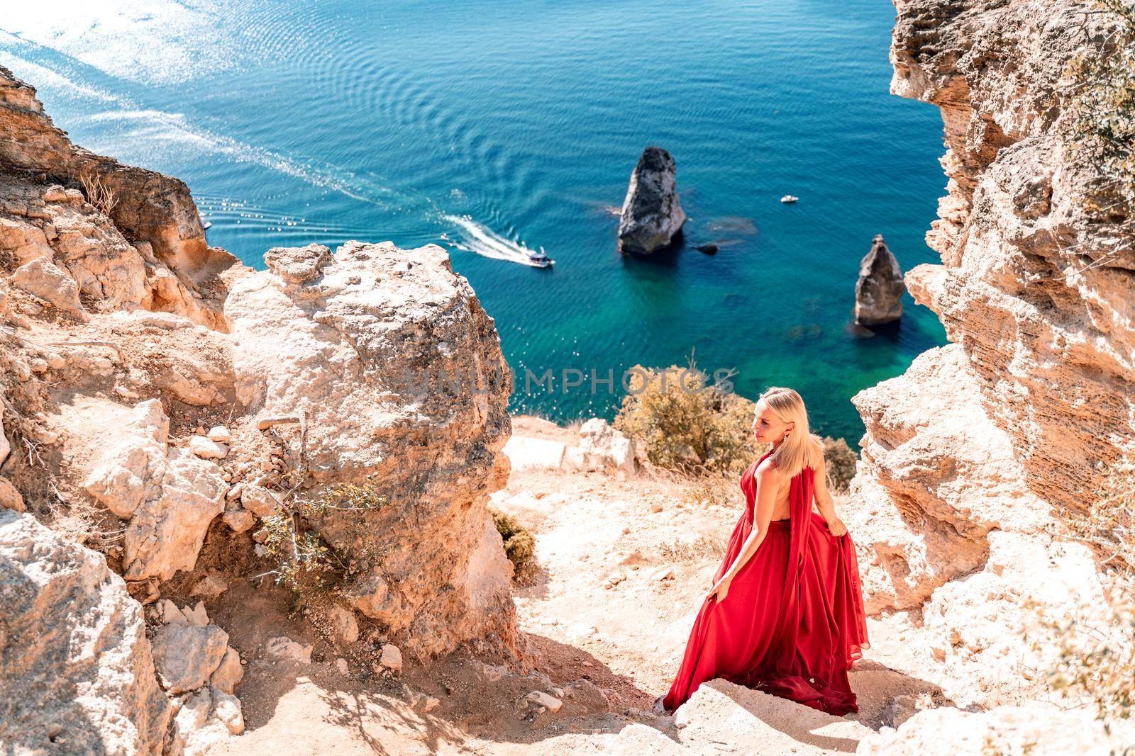 A girl with loose hair in a long red dress descends the stairs between the yellow rocks overlooking the sea. A rock can be seen in the sea. Sunny path on the sea from the rising sun.