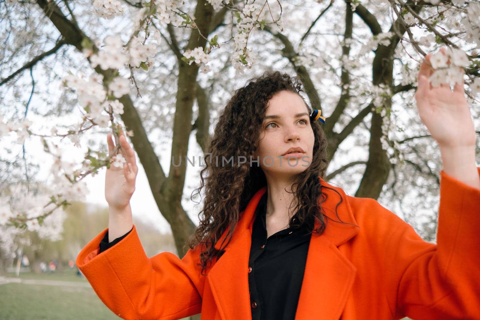 portrait of curly brunette woman in red coat in the park near flowering tree with ukrainian symbol in hair