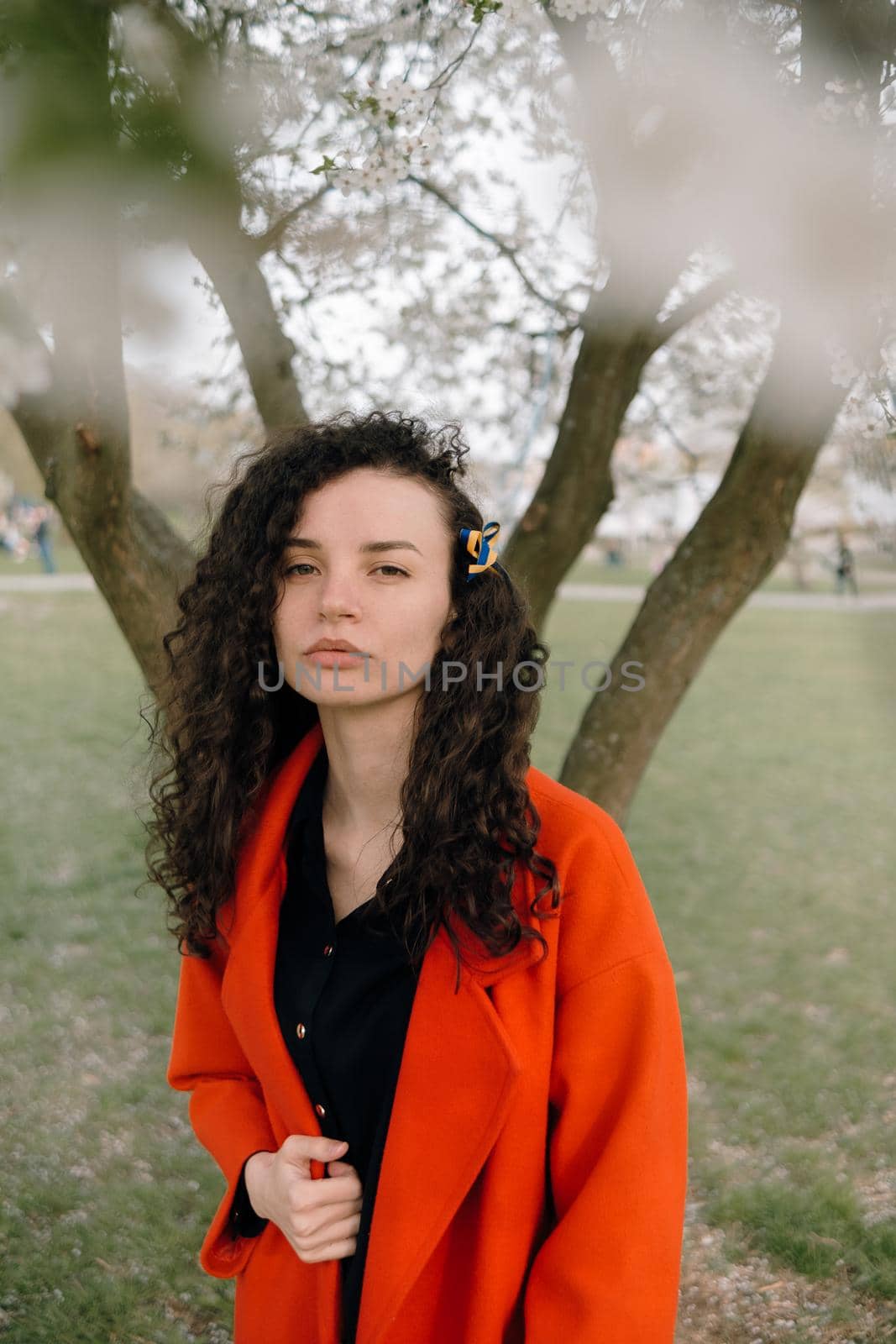 portrait of curly brunette woman in red coat in the park near flowering tree with ukrainian symbol in hair