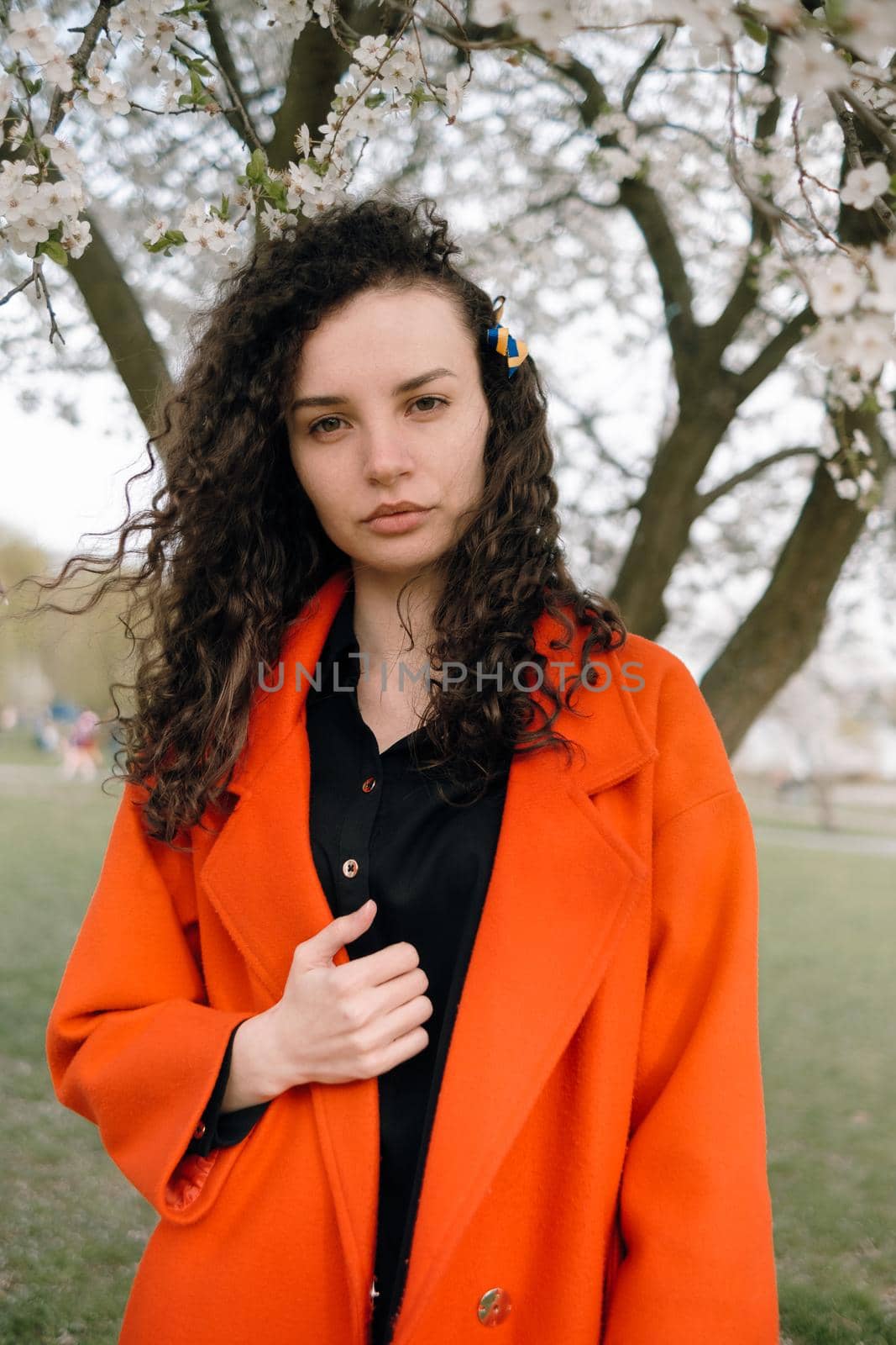 portrait of curly brunette woman in red coat in the park near flowering tree with ukrainian symbol in hair by Symonenko