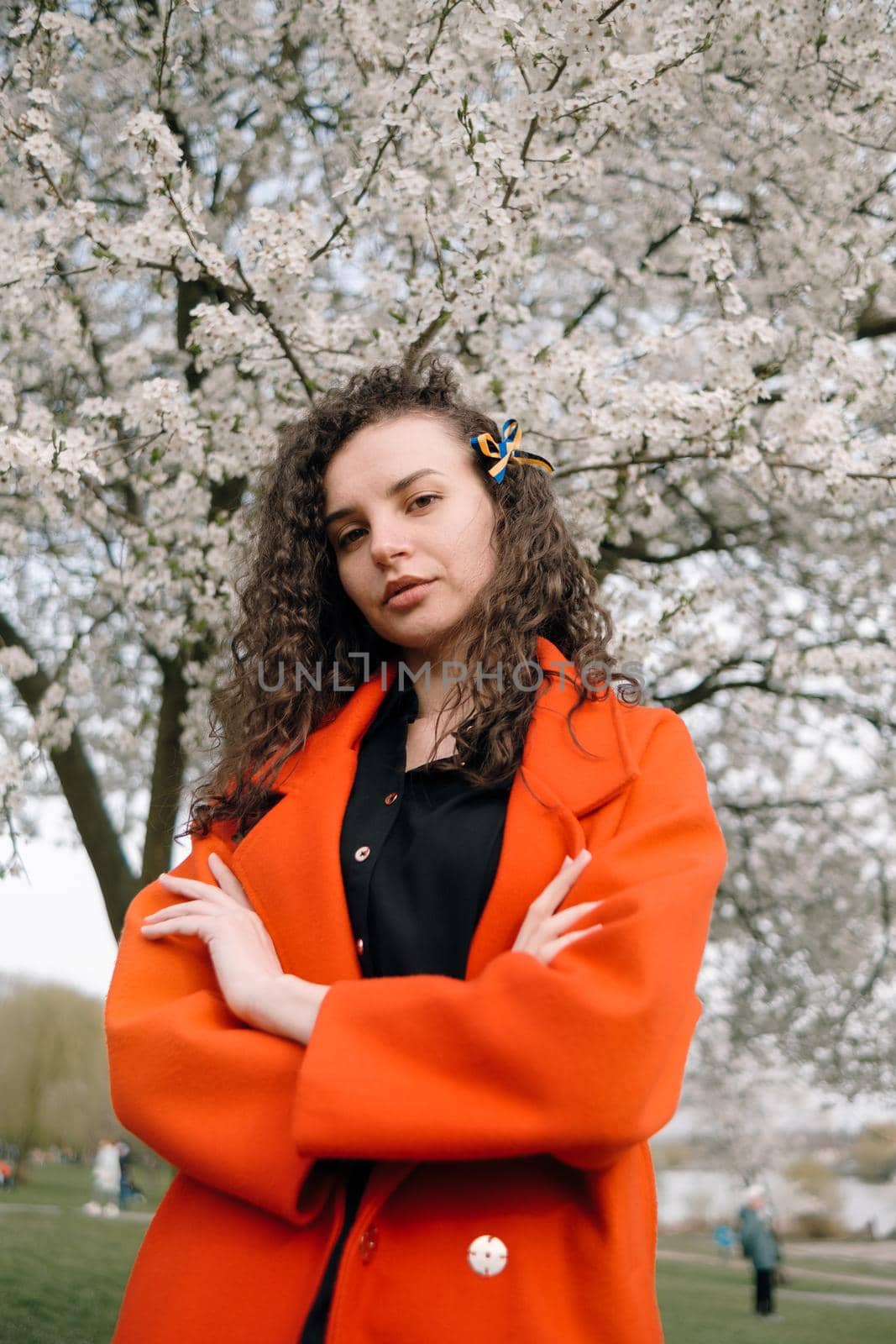 portrait of curly brunette woman in red coat in the park near flowering tree with ukrainian symbol in hair