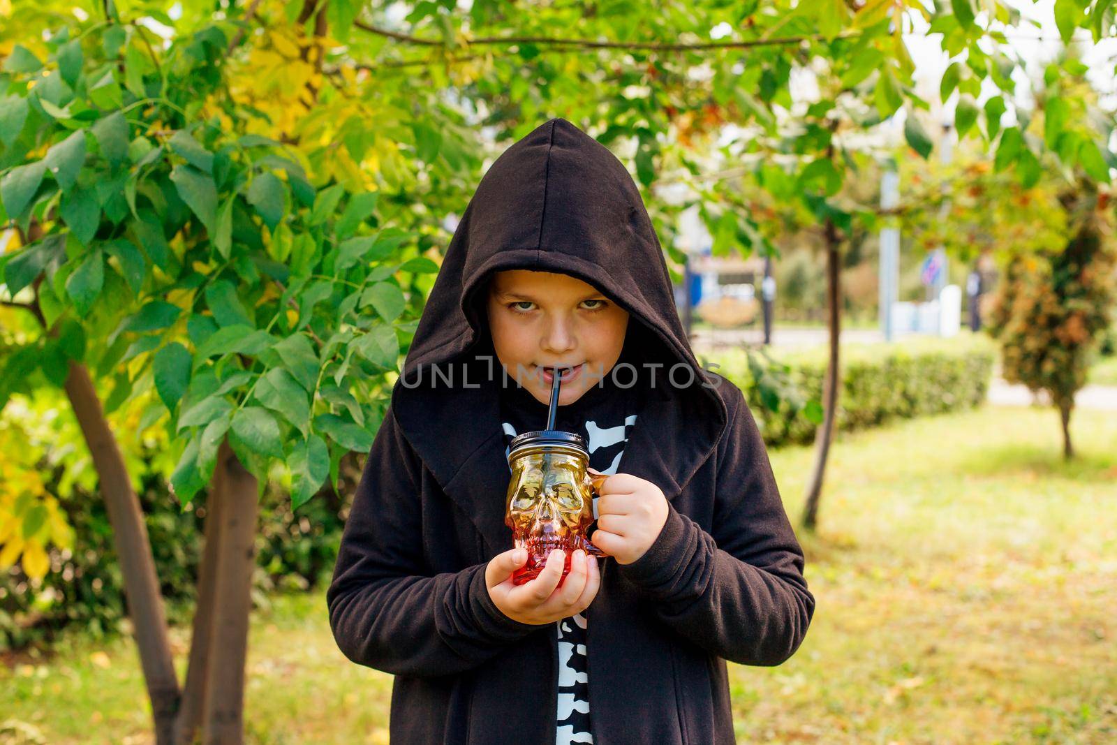 Smiling boy in black costume of skeleton and hood holding glass in the shape of a skull outdoors.