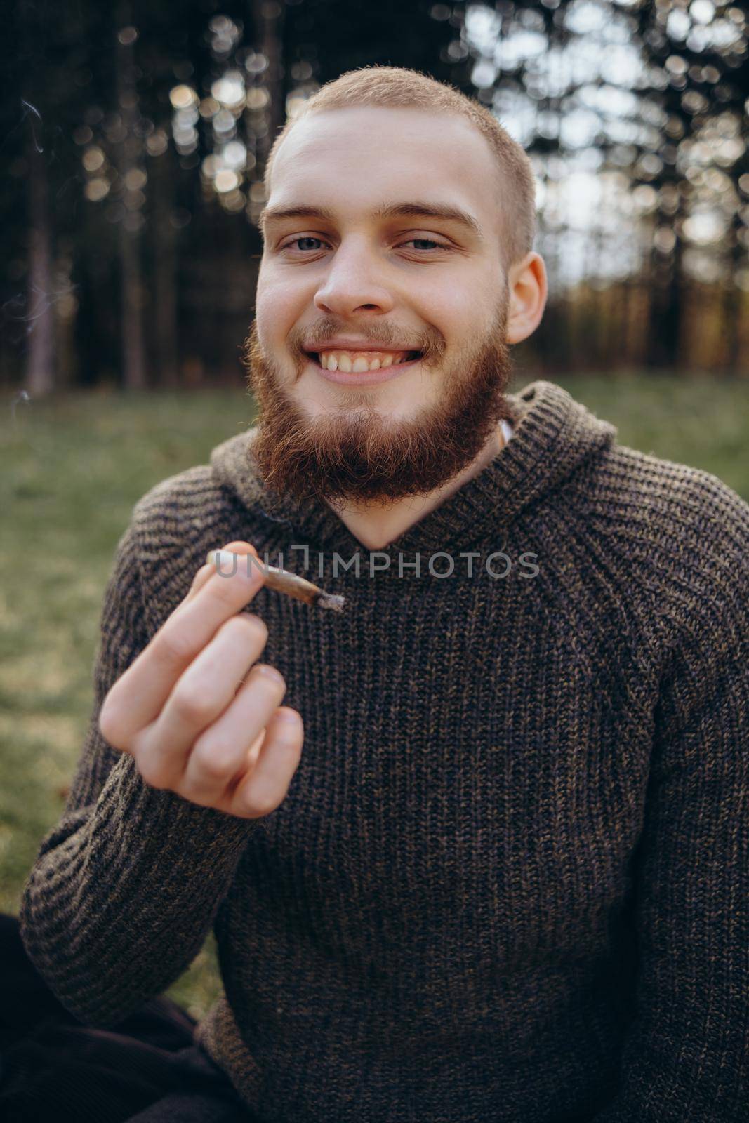 Young man beard smokes cigarette or tobacco. Guy is addicted to cigarettes and always smokes lot of tobacco every day. He smokes cigarette outdoors on background of lake in park. Cool guy blows smoke
