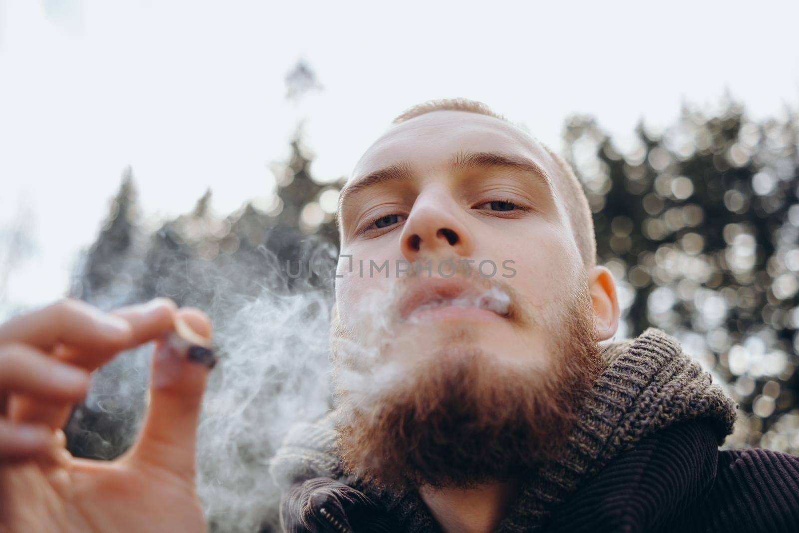 Young man beard smokes cigarette or tobacco. Guy is addicted to cigarettes and always smokes lot of tobacco every day. He smokes cigarette outdoors on background of lake in park. Cool guy blows smoke