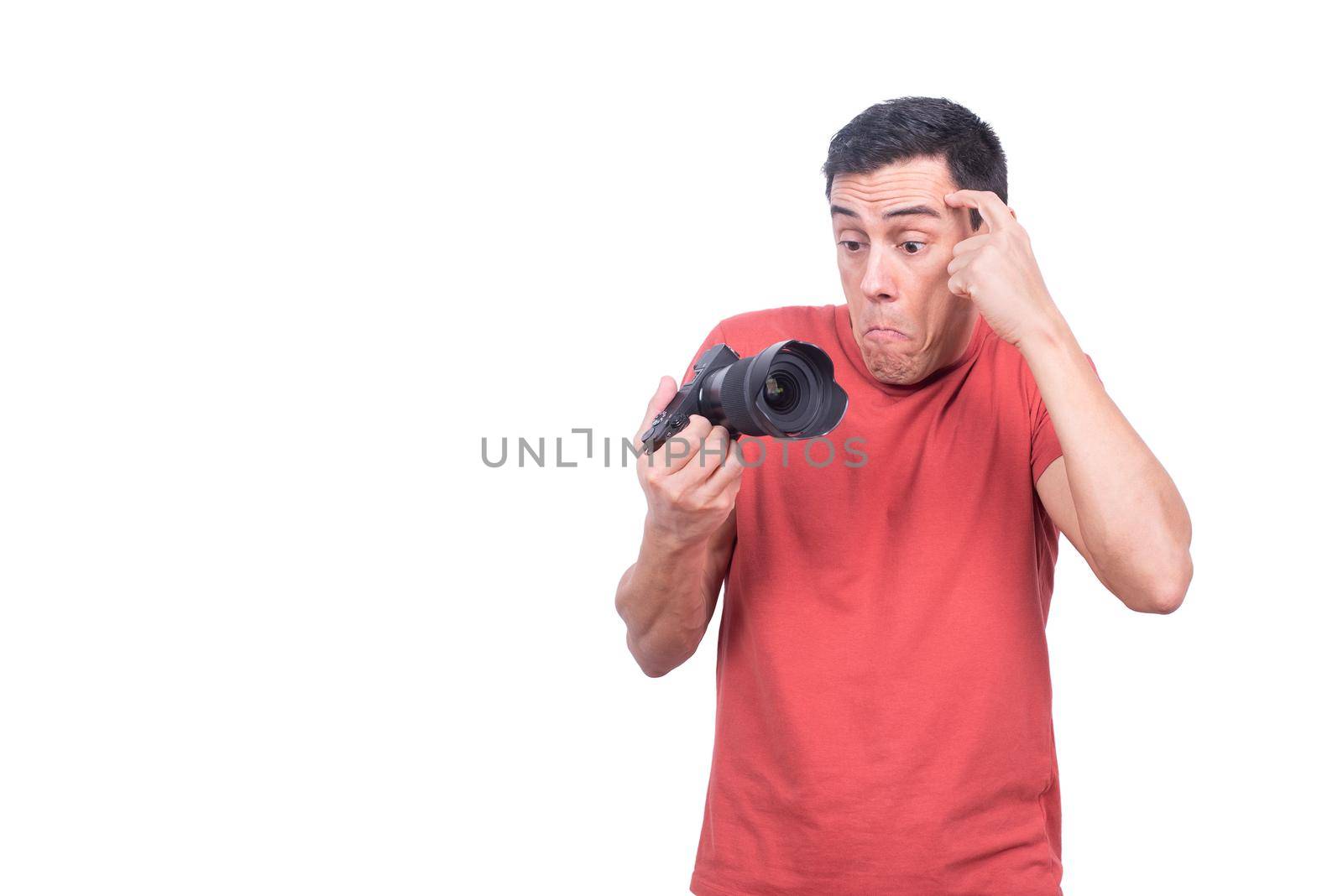 Confused novice male photographer looking at professional photo camera with perplexity while standing isolated on white background in light studio
