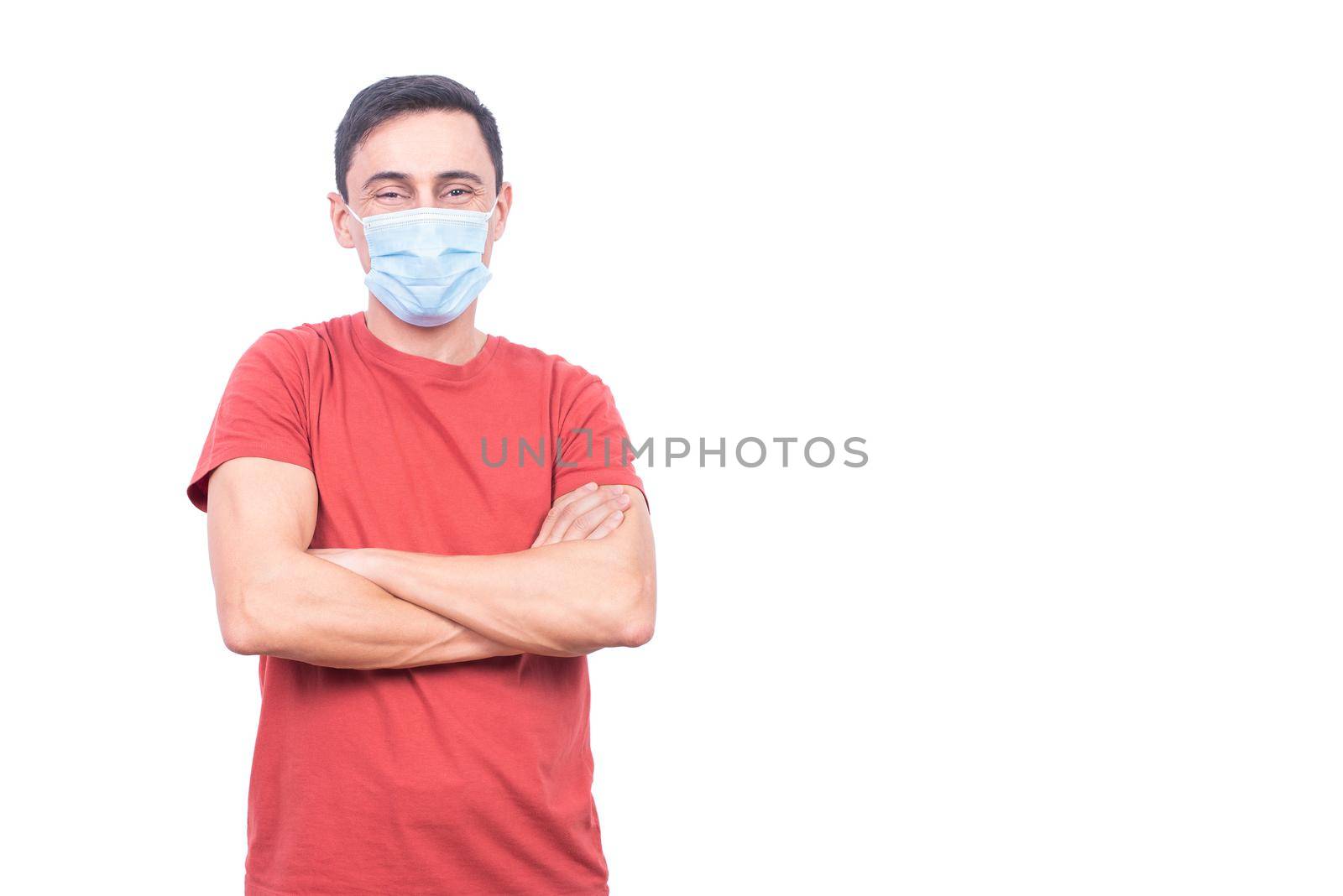 Optimistic male in protective mask looking at camera with crossed arms while standing isolated on white background in light studio