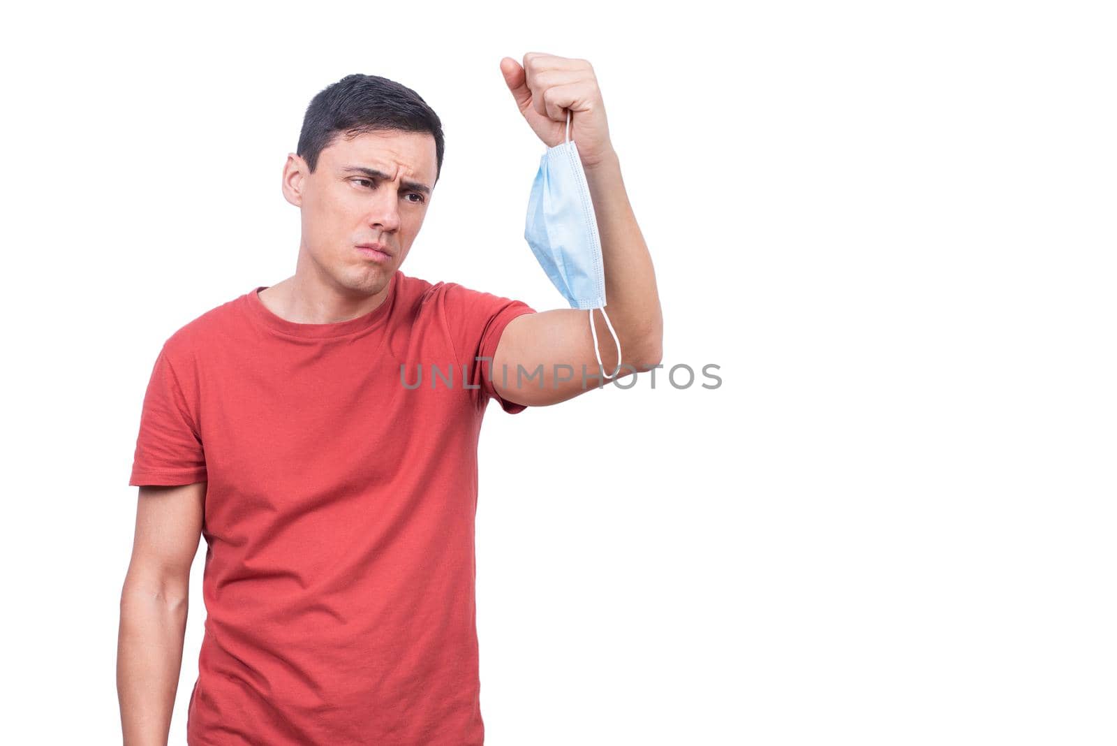 Sad male looking at protective mask in hand while standing isolated on white background during coronavirus pandemic in light studio