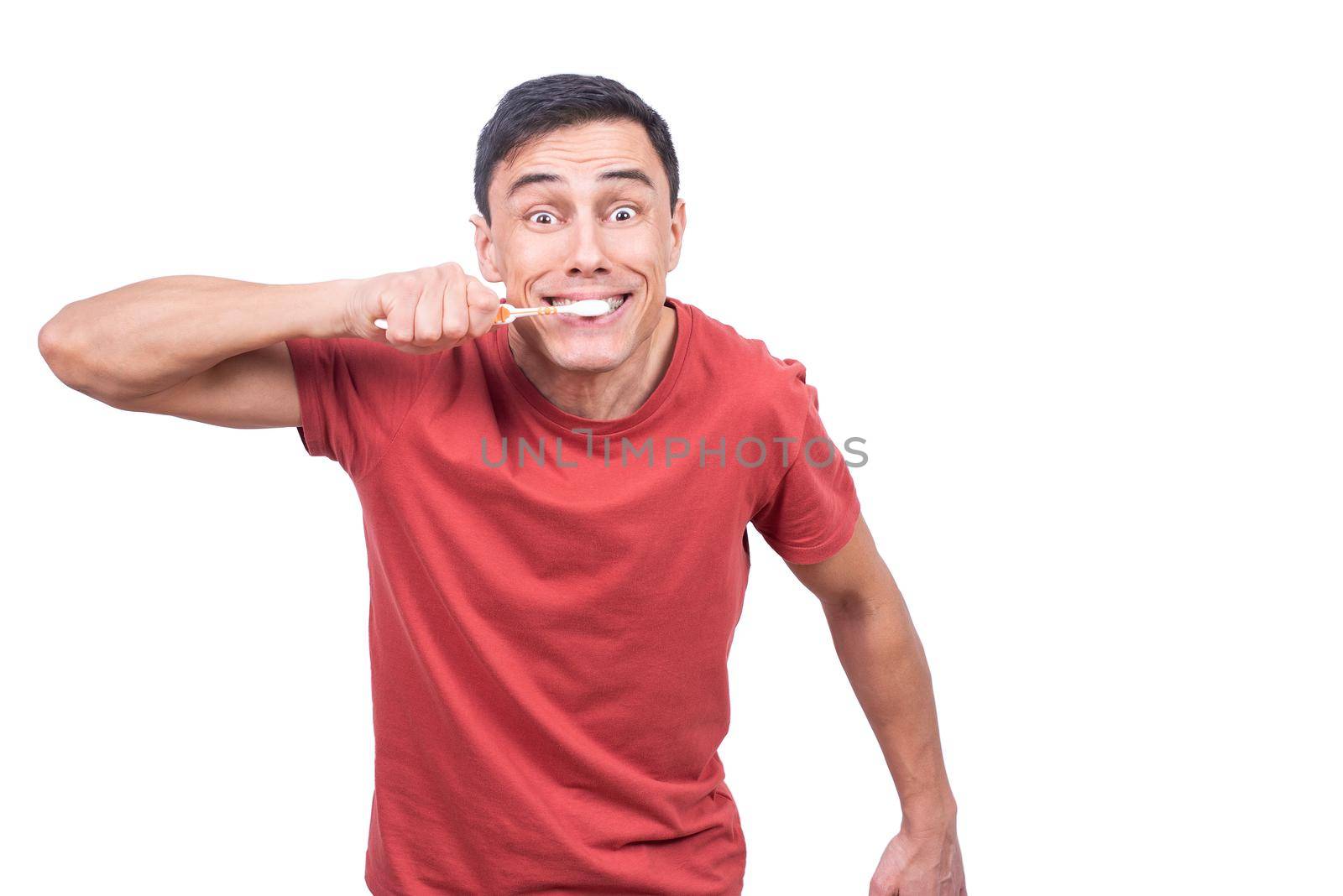 Male in red t shirt looking at camera while standing on white background and brushing teeth in studio