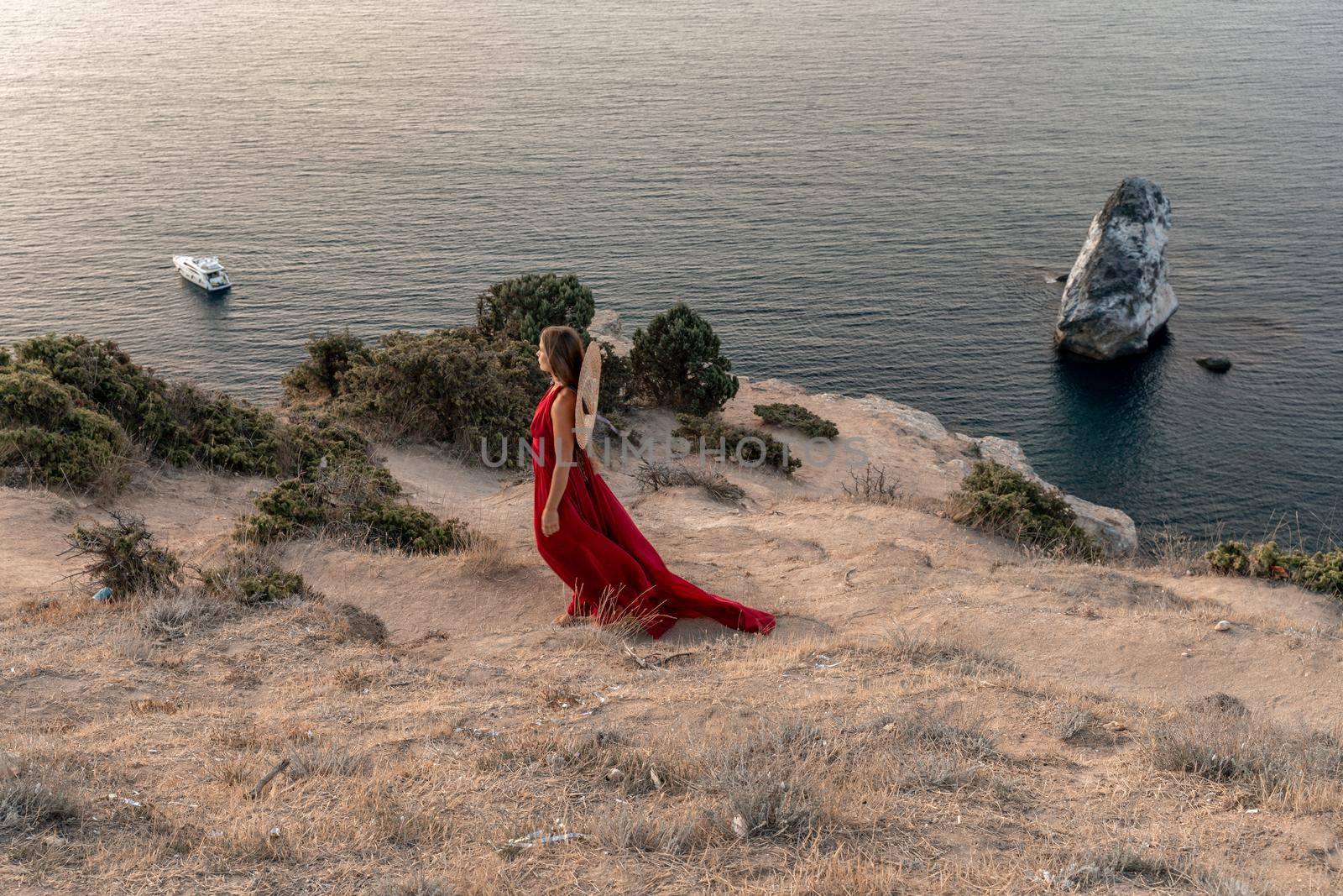 A girl with loose hair in a red dress and hat stands on the rocks above the sea. In the background, the sea and the rocks. The concept of travel. by Matiunina
