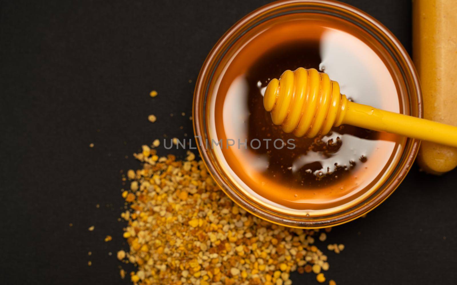 A yellow spoon of honey dipped in a jar of honey and bee bread scattered on a black background. Honey dripping around, nice and inviting photos. Healthy food concept. by Matiunina