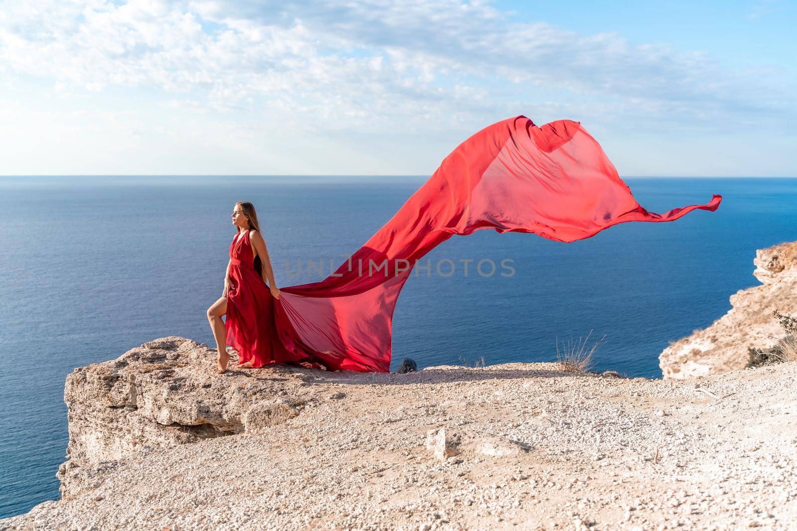 A girl with loose hair in a red dress stands on a rock rock above the sea. In the background, the sea and the rocks. The concept of travel by Matiunina