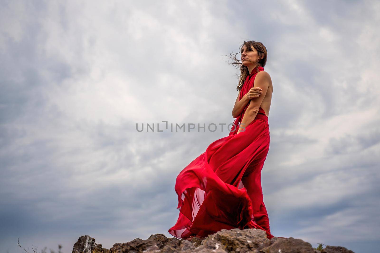 A woman in a red dress stands above a stormy sky, her dress fluttering, the fabric flying in the wind