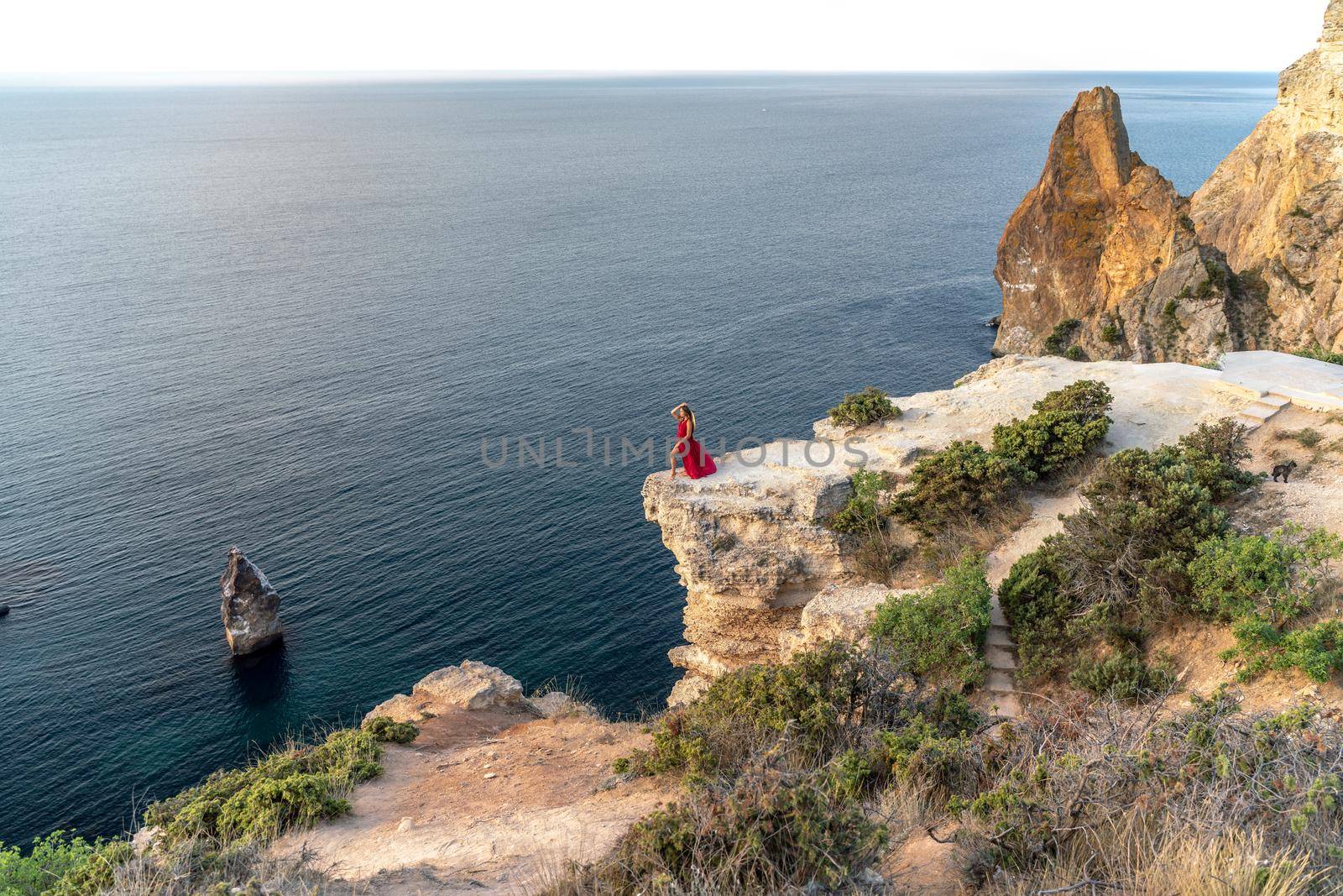 A girl with loose hair in a red dress stands on a rock rock above the sea. In the background, the sea and the rocks. The concept of travel.