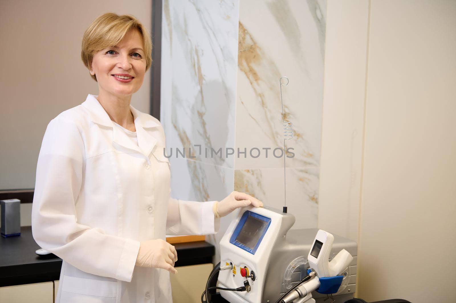 Experienced charming Caucasian mature female doctor in white medical gown and sterile surgical gloves , smiles at camera standing near ultrasound medical equipment in doctor's cabinet
