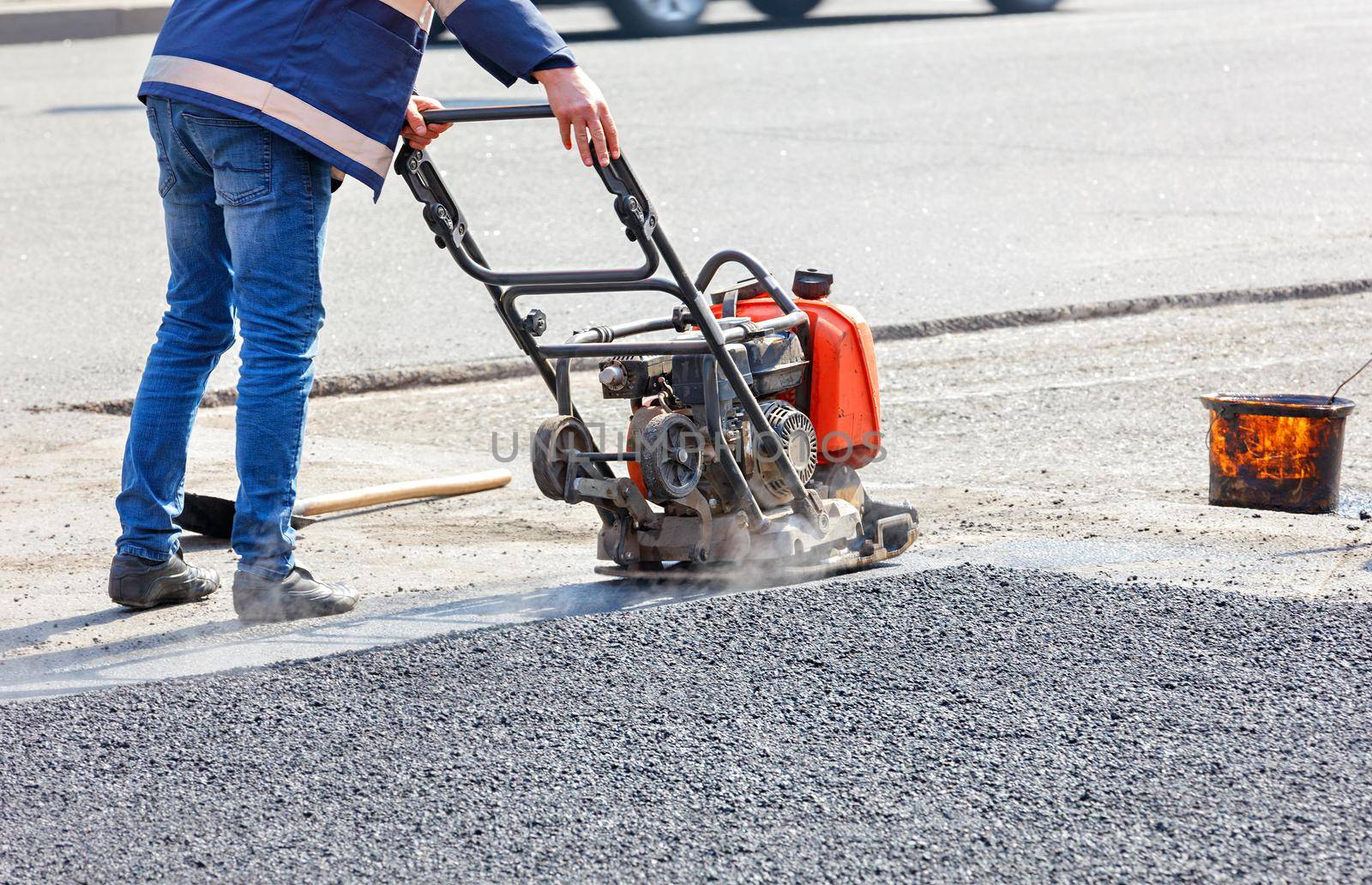 A road service worker compacts the asphalt on a fenced road section of the roadway with a petrol vibration plate compactor.
