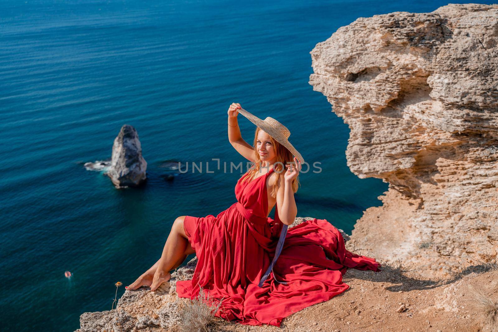 A girl with flowing hair in a long red dress sits on a rock above the sea. The stone can be seen in the sea. by Matiunina