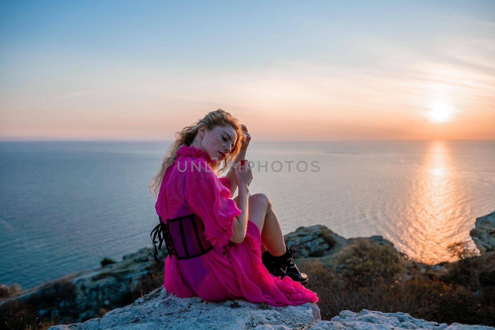 Side view a Young beautiful sensual woman in a red long dress posing on a volcanic rock high above the sea during sunset. Girl on the nature on overcast sky background. Fashion photo