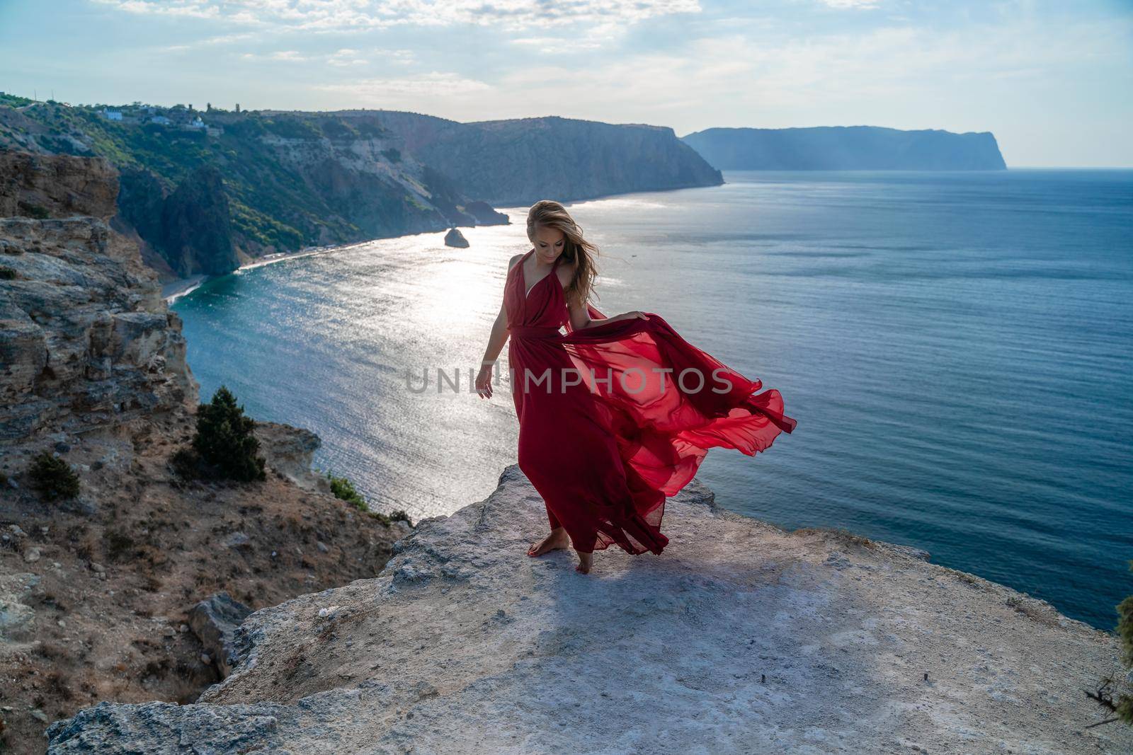 A girl with flowing hair in a long red dress stands on a rock above the sea. The stone can be seen in the sea. Sunny path to the sea from the sun. by Matiunina