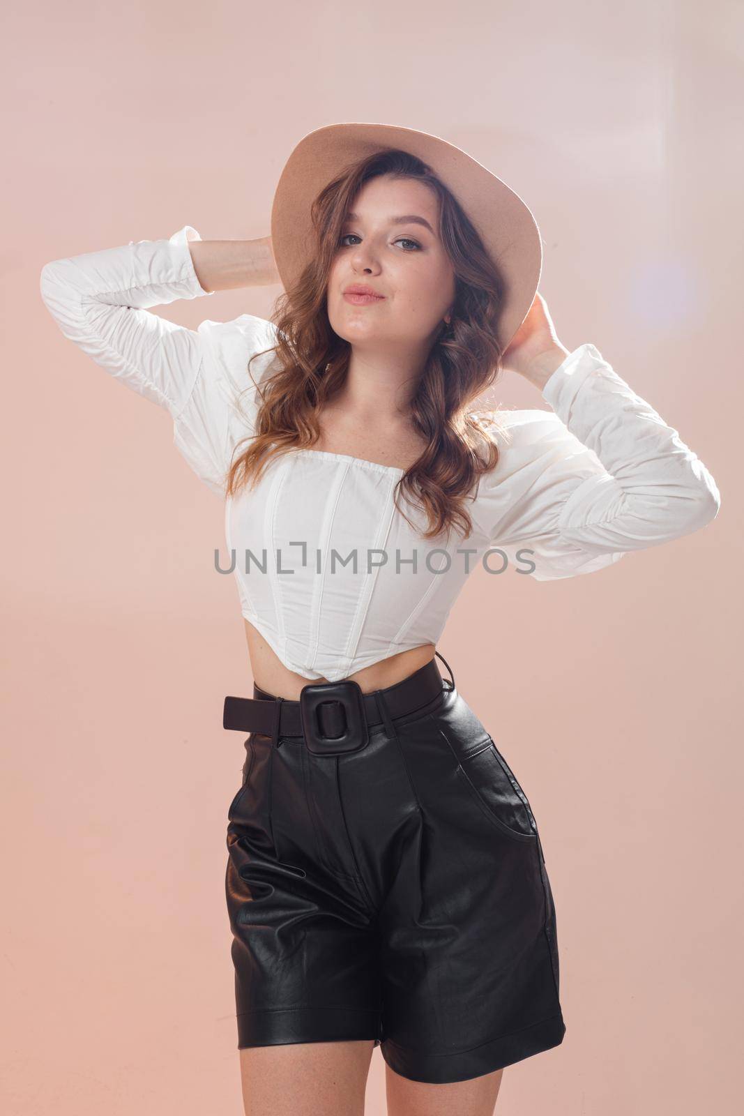 A beautiful beautiful woman in a white bodysuit shirt and leather shorts, a hat on her head, in the studio on the background. by StudioLucky