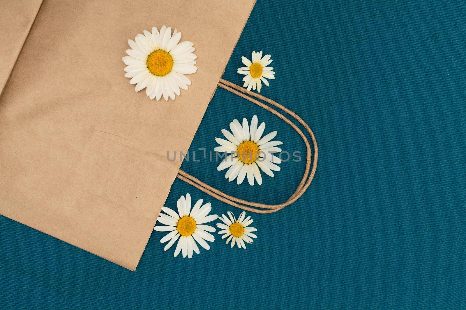 Paper bag on a blue background, decorated with daisies, brown shopping bag. Craft bag with handle. Packaging template mockup. Delivery service concept. Copy space.