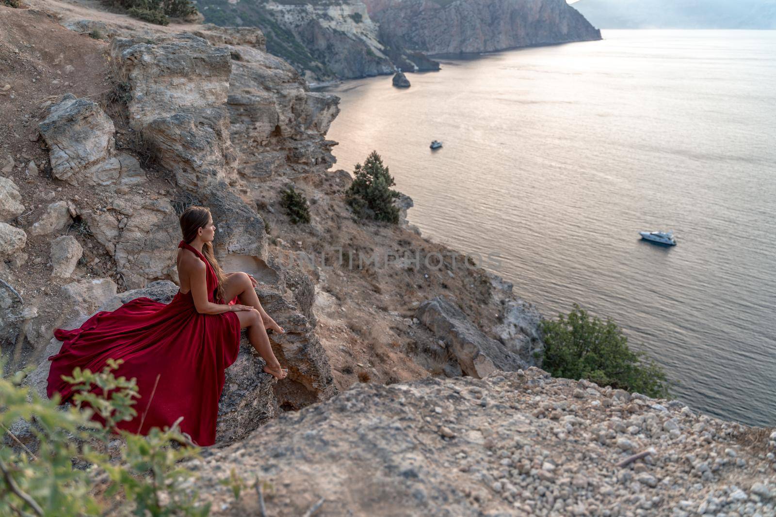 A girl with loose hair in a red dress sits on a rock rock above the sea. In the background, the sea. The concept of travel