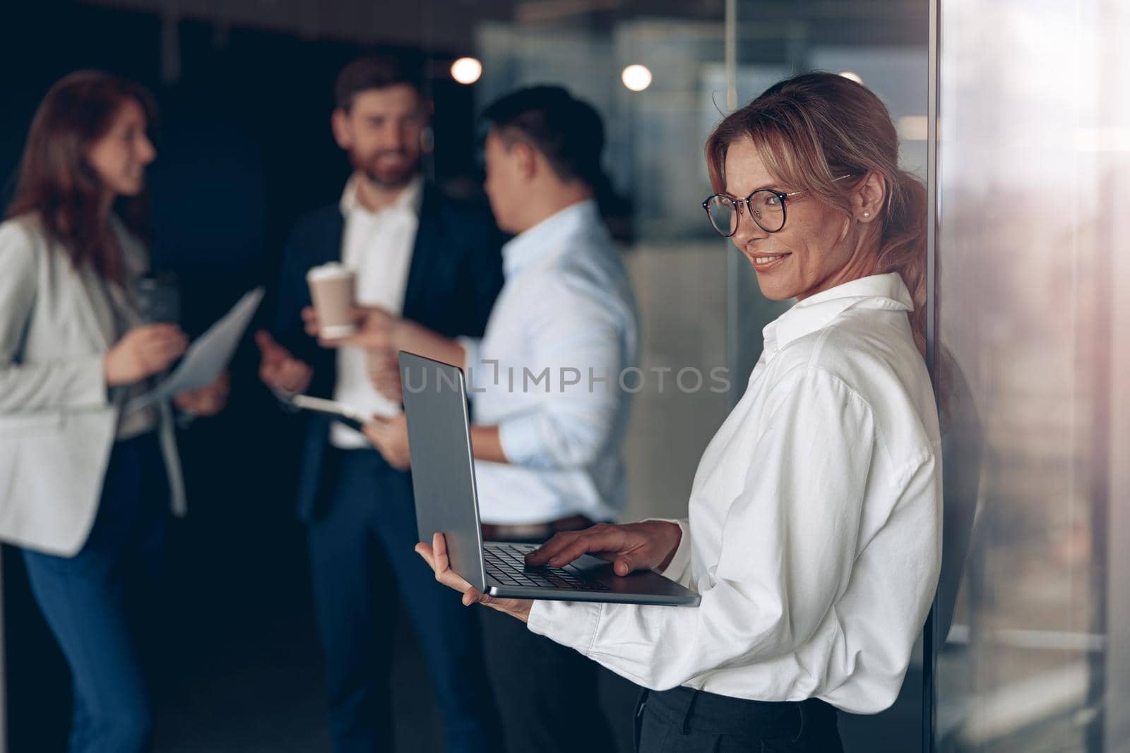 Mature smiling business lady at office with group of colleagues on background, working on laptop.