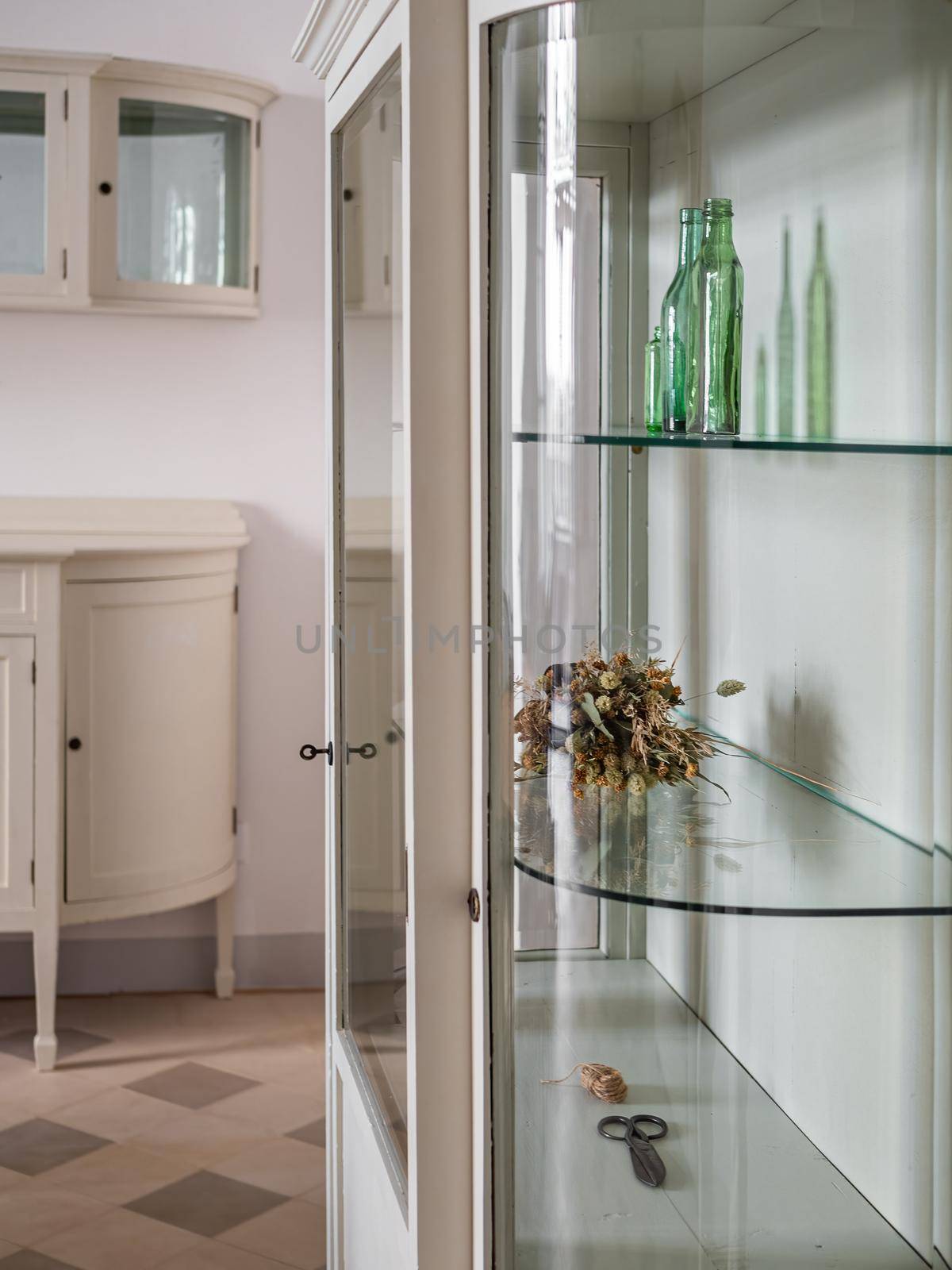 White old style cabinets with glass shelves in a living room. Green glass bottles and dried flowers for decoration