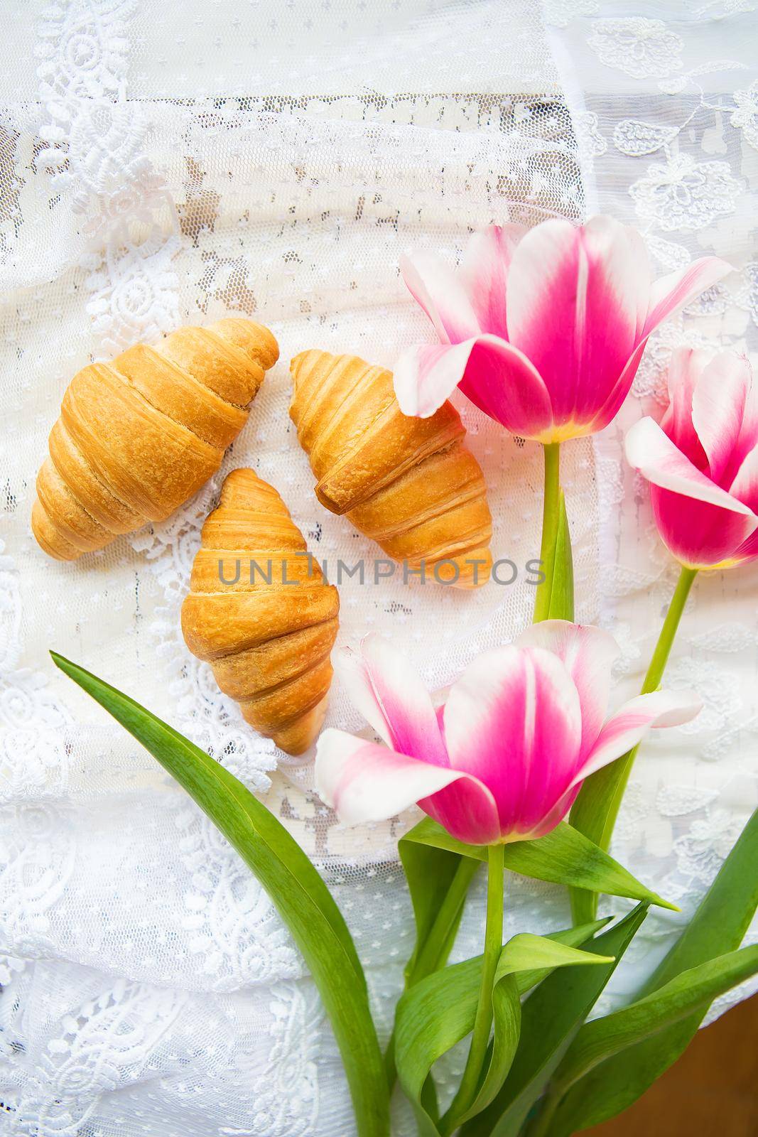 Three croissants and bright pink tulips on lace tablecloth.