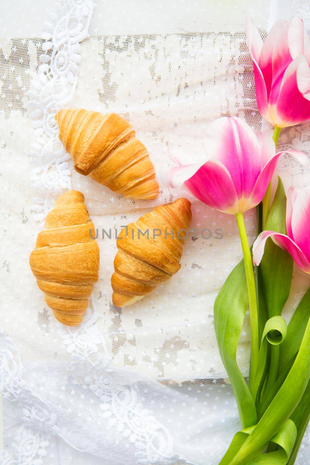 Three croissants and bright pink tulips on lace tablecloth, close-up.