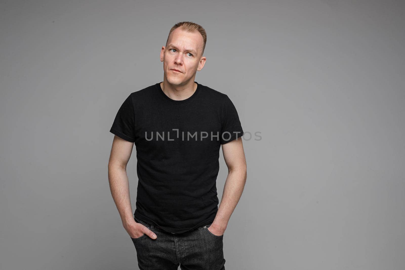 picture of caucasian man keeps his hands in the pockets of his black jeans and looks to the right isolated on grey background
