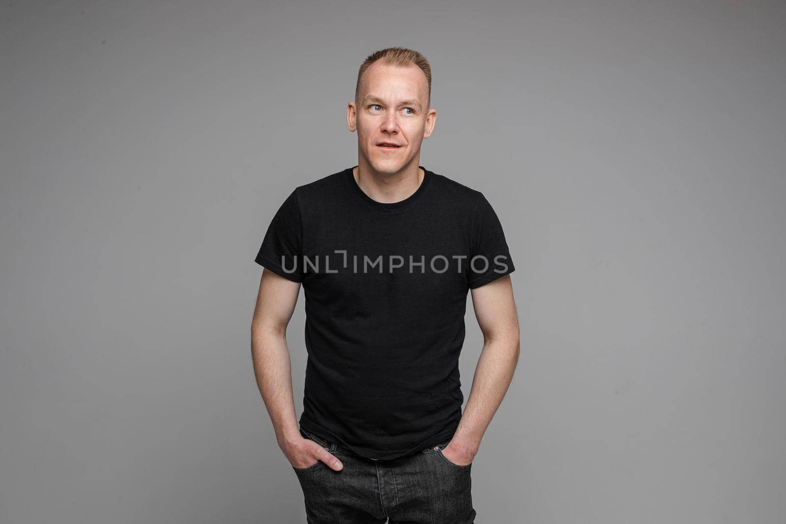 picture of caucasian man keeps his hands in the pockets of his black jeans and looks to the right isolated on grey background