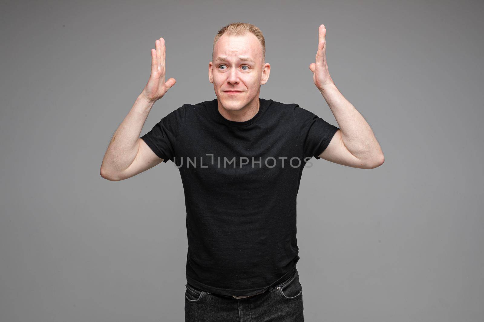 Portrait of puzzled blond Caucasian man in black t-shirt holding arms up showing misunderstanding and disbelief. He doesn’t understand what is happening.