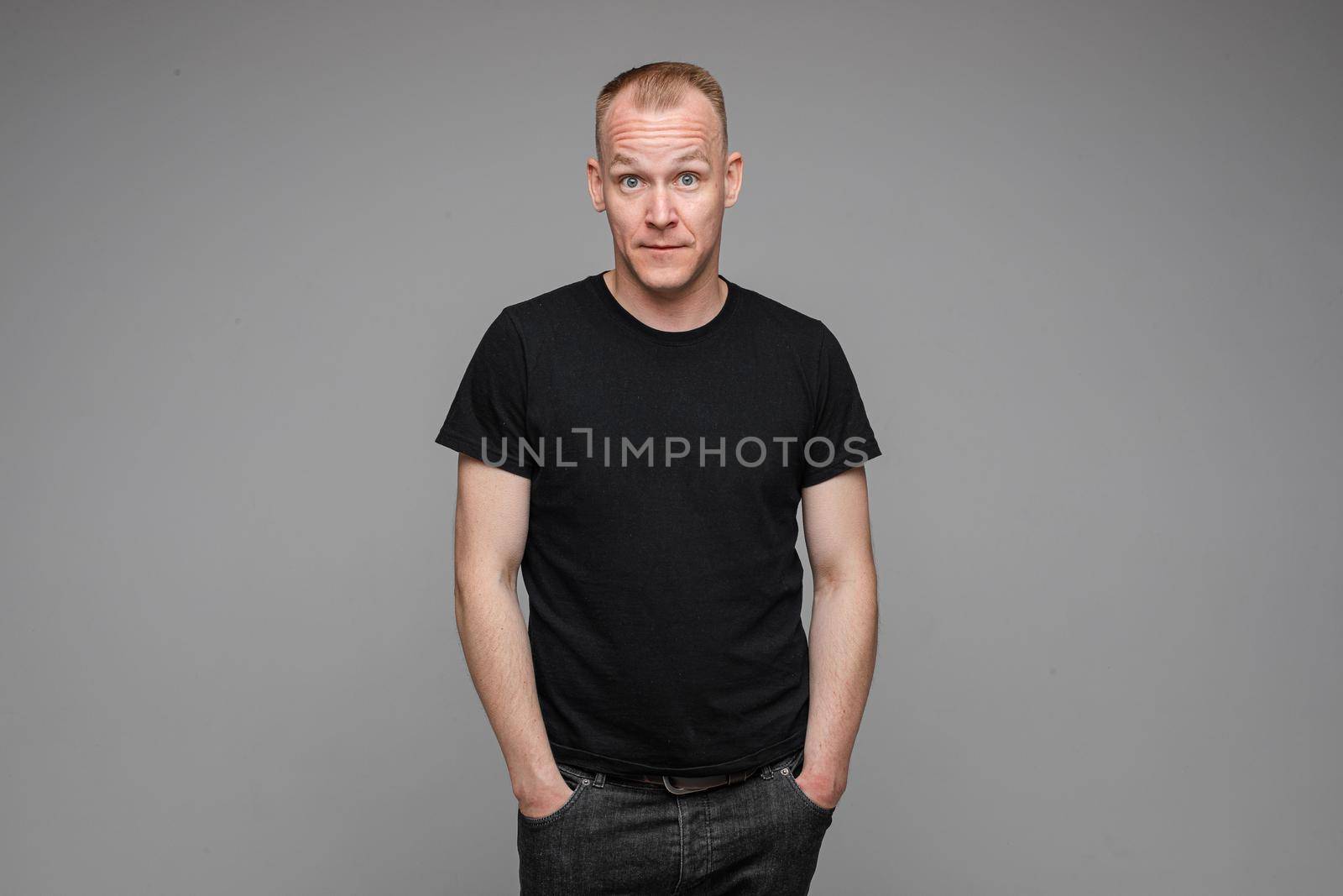 Portrait of a man in black looking at camera with amazed wide open eyes holding hands in pockets of his black jeans. Isolate on grey background.