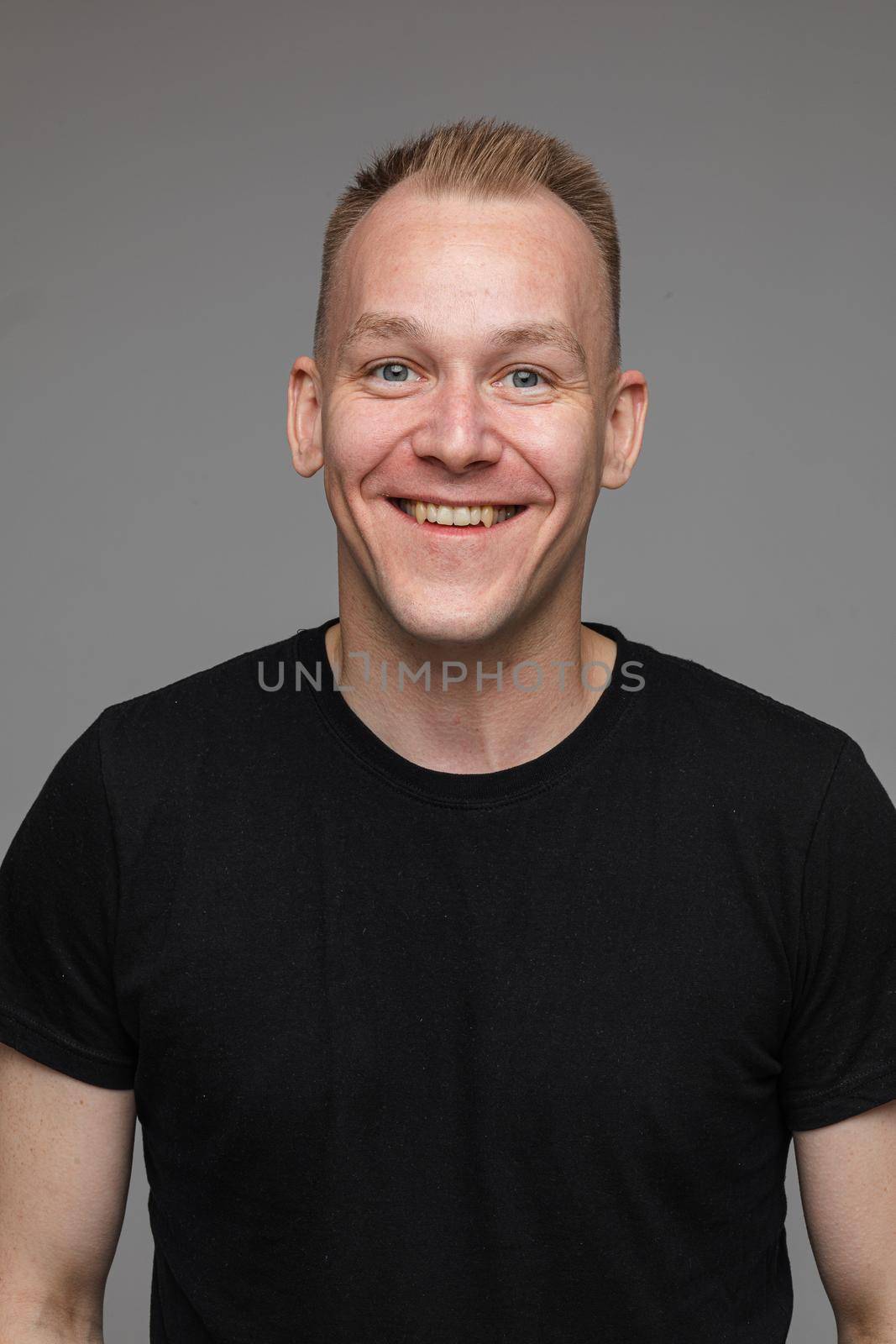 Studio portrait of adult man with short blond hair wearing casual black t-shirt smiling cheerfully at camera with friendly look. Cutout on grey background.