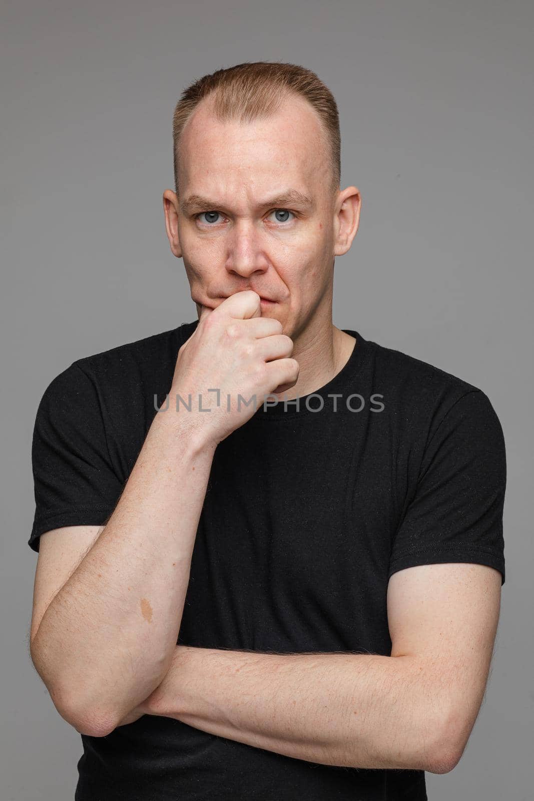 portrait of caucasian man in black t-shirt looks to the camera and keeps his hand near the face isolated on grey background by StudioLucky