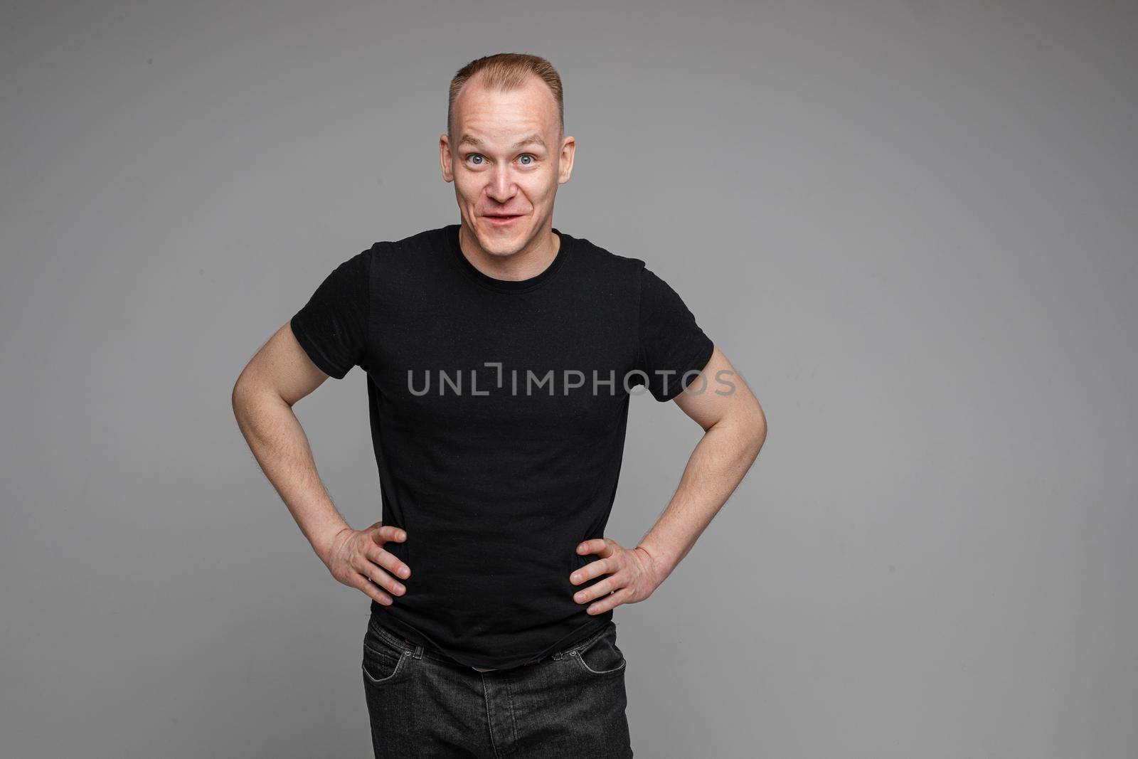 Surprised Caucasian man in black looking at camera with smile with hands on his waist.