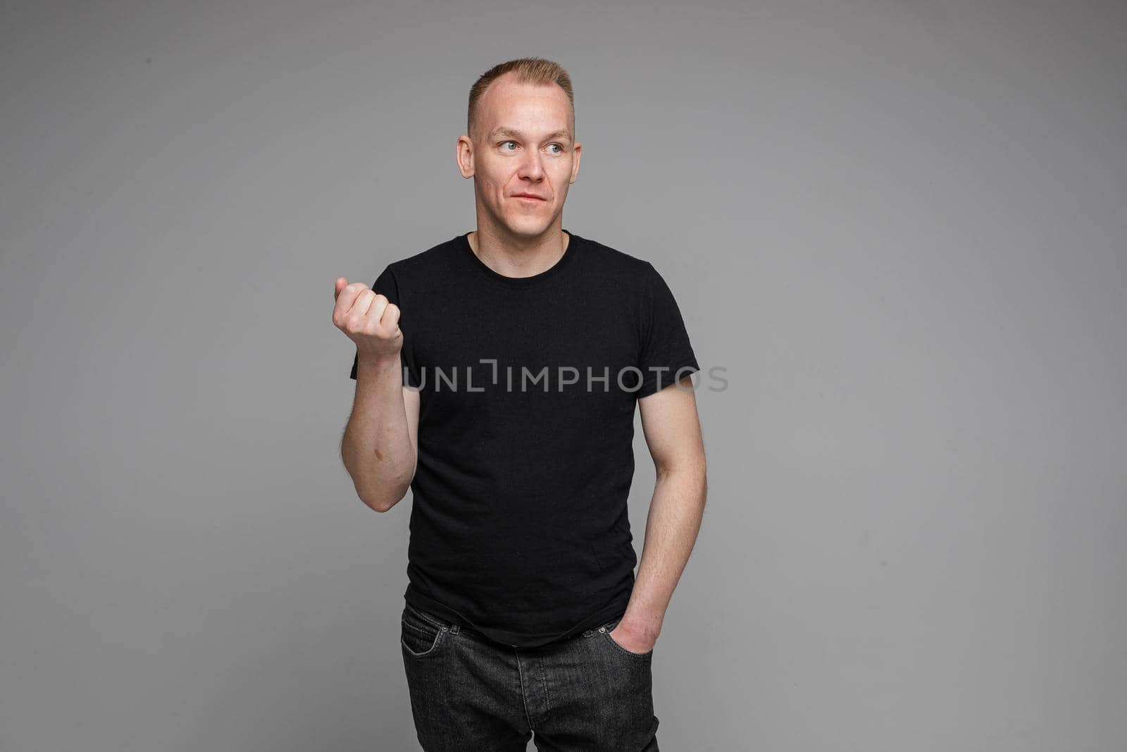 picture of caucasian man keeps his hands in the pockets of his black jeans and looks to the right isolated on grey background