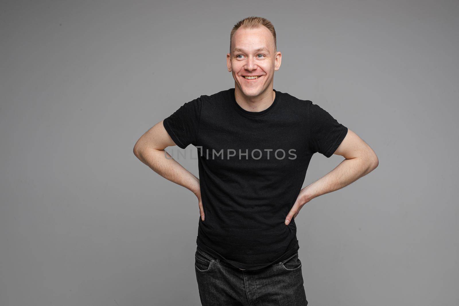 Portrait of blond Caucasian man in black t-shirt and dark jeans holding hands together with eyes closed on grey background. Bored or tired asleep man standing on grey background.