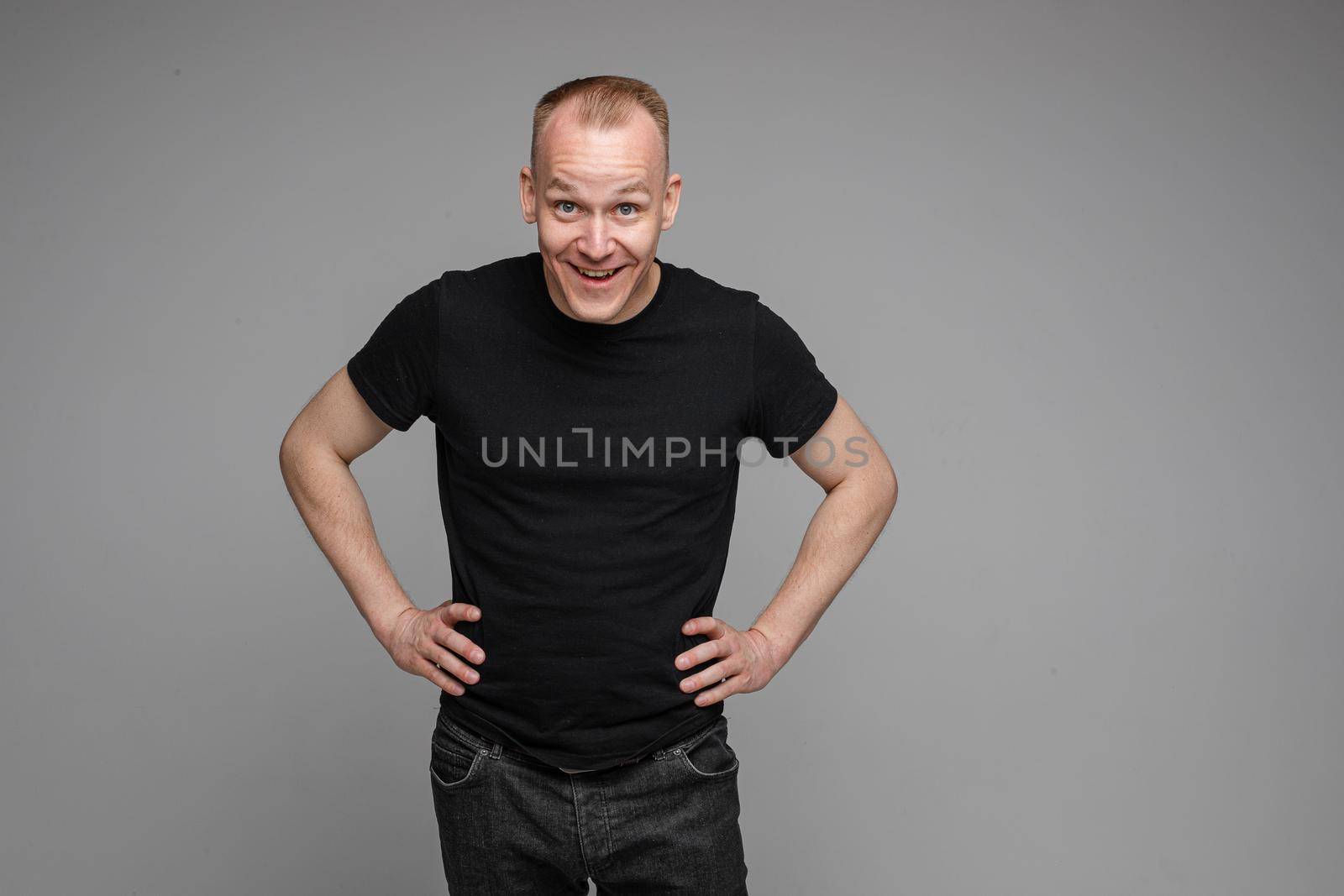 Surprised Caucasian man in black looking at camera with smile with hands on his waist.