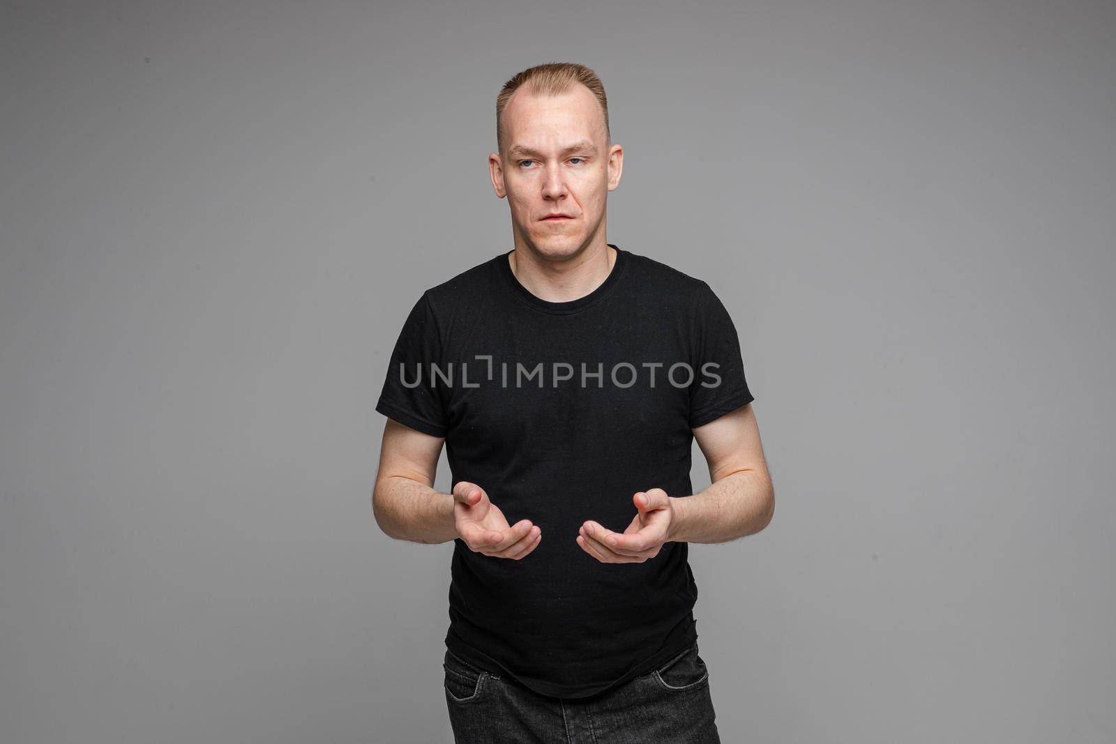 Portrait of blond Caucasian man in black t-shirt and dark jeans holding hands together with eyes closed on grey background. Bored or tired asleep man standing on grey background.
