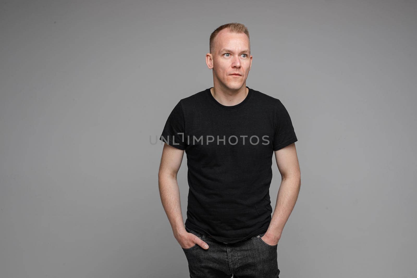 picture of caucasian man keeps his hands in the pockets of his black jeans and looks to the right isolated on grey background