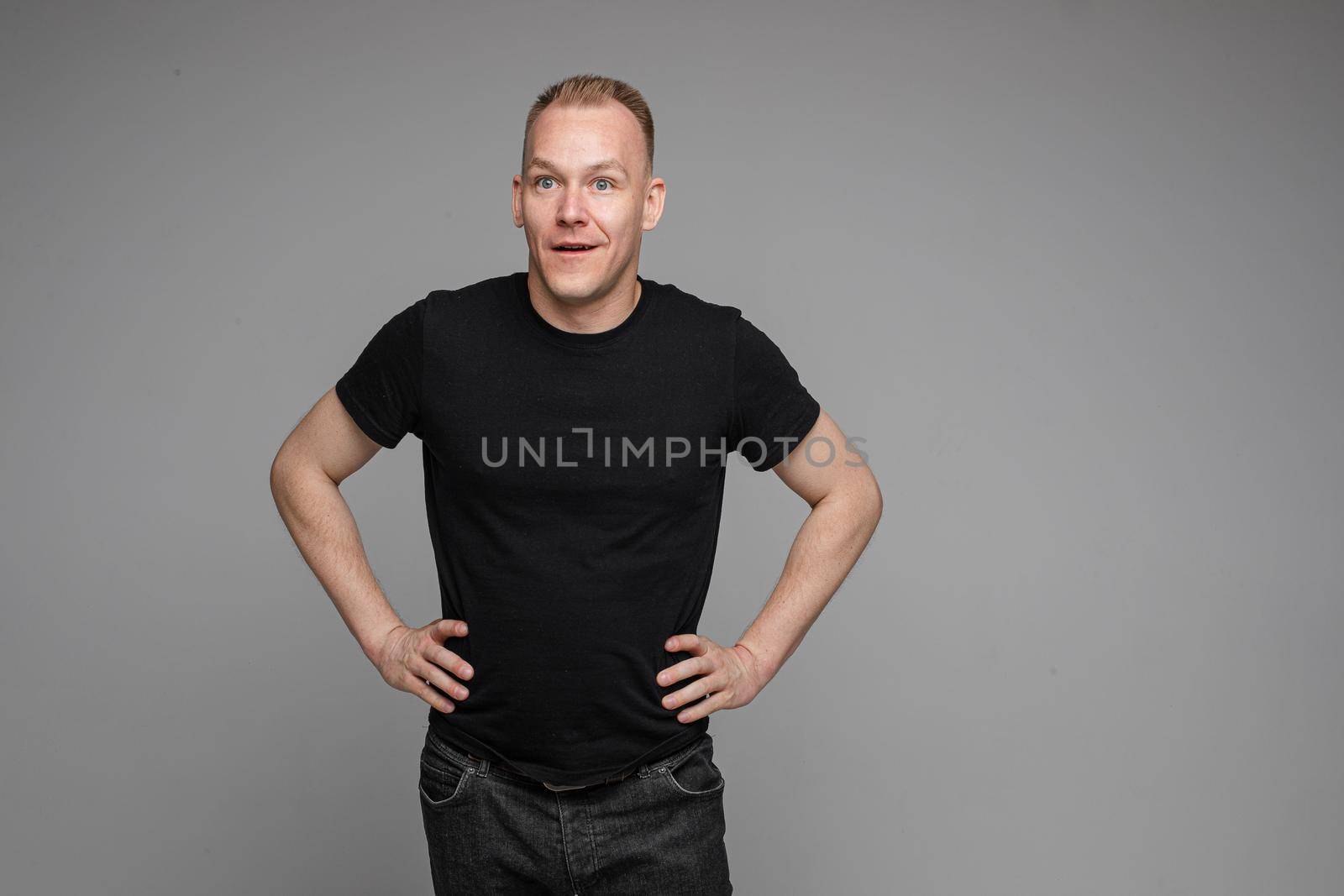 Surprised Caucasian man in black looking at camera with smile with hands on his waist.