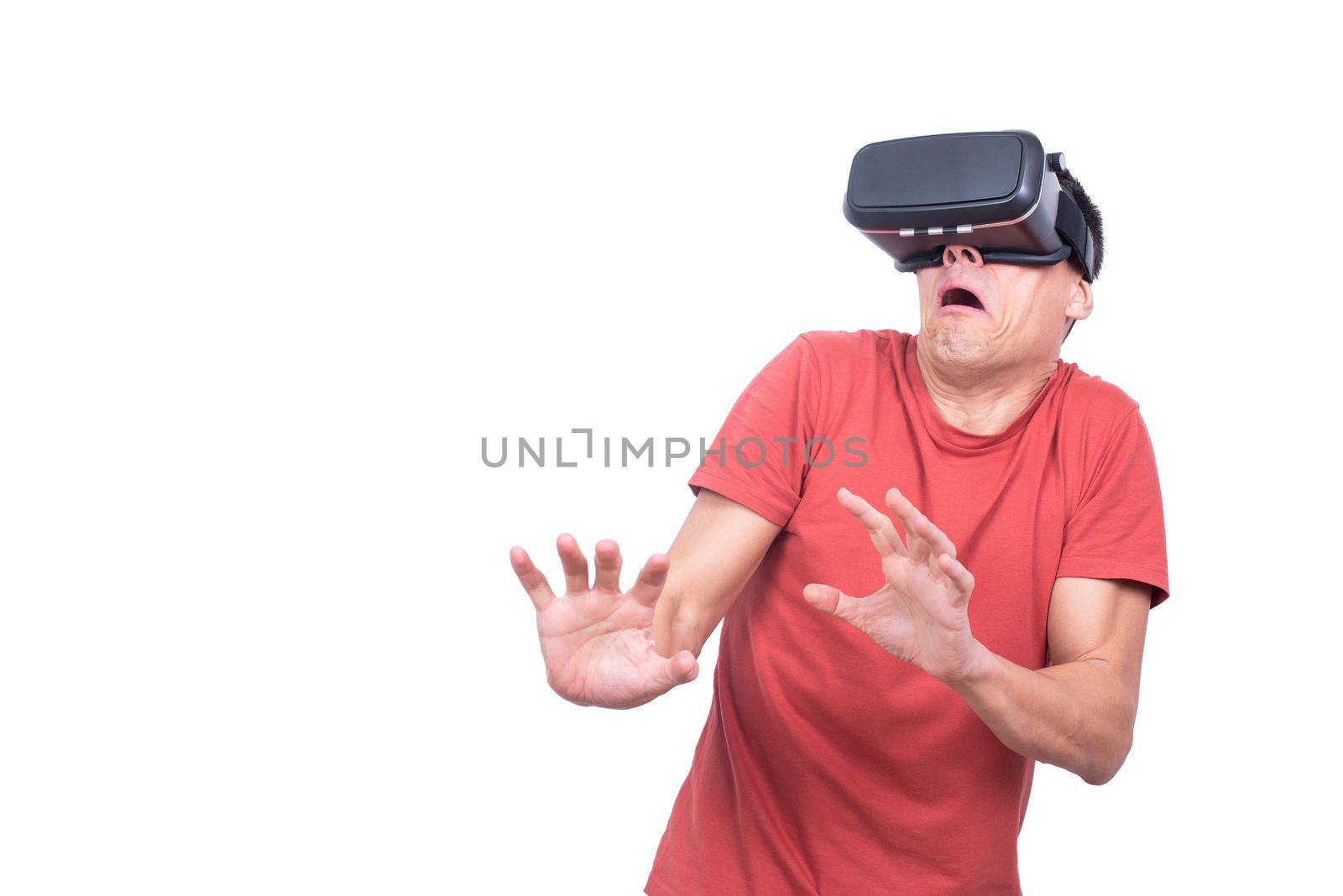Fearful male in red t shirt and modern VR goggles exploring virtual reality while standing on white background in light studio