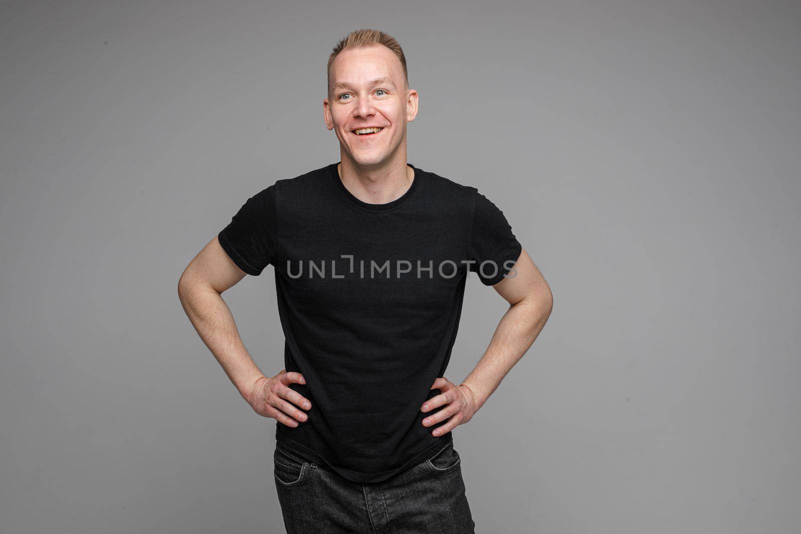 Portrait of serious blond Caucasian man in black t-shirt and dark jeans holding hands with fingers crossed looking attentively away at someone. Isolate on grey background.