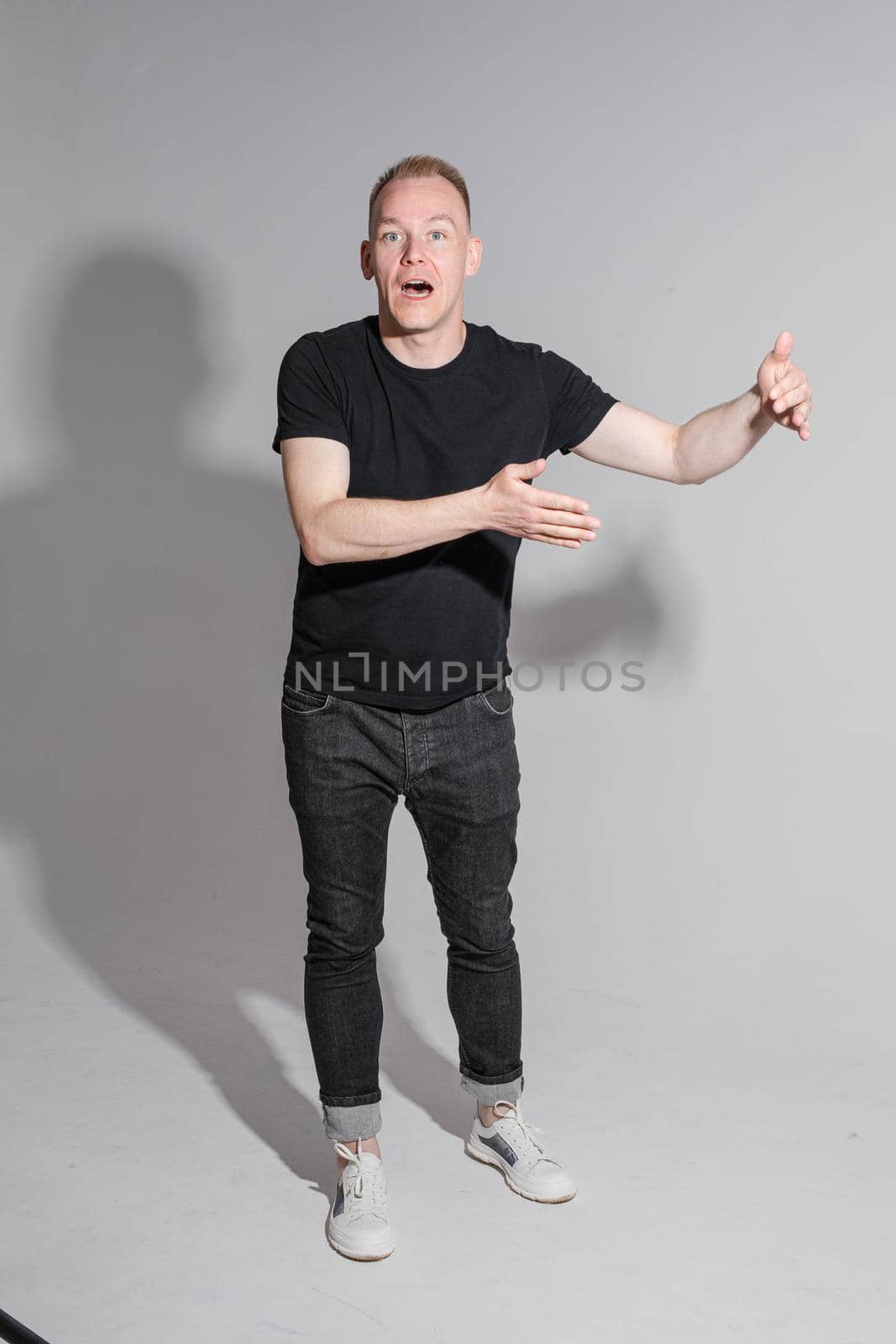 Full length of adult Caucasian blonde man in black t-shirt, dark jeans and sneakers talking to camera with arms bent, holding fists. Presenter or advertiser promoting something.